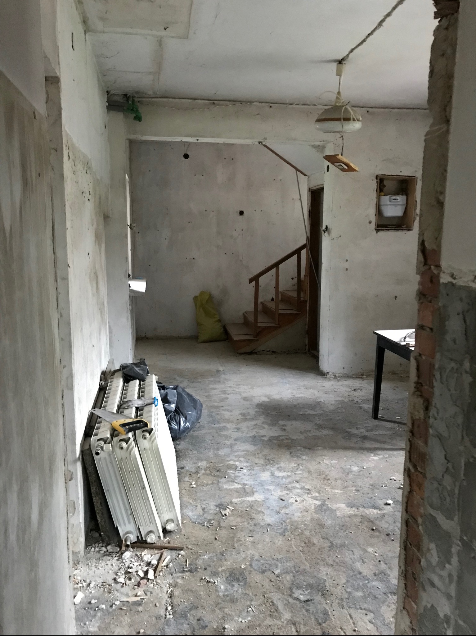 Wnętrze w trakcie remontu: surowe ściany, demontowane grzejniki oparte o ścianę, drewniane schody w tle, widoczny gruz na podłodze i wisząca lampa.