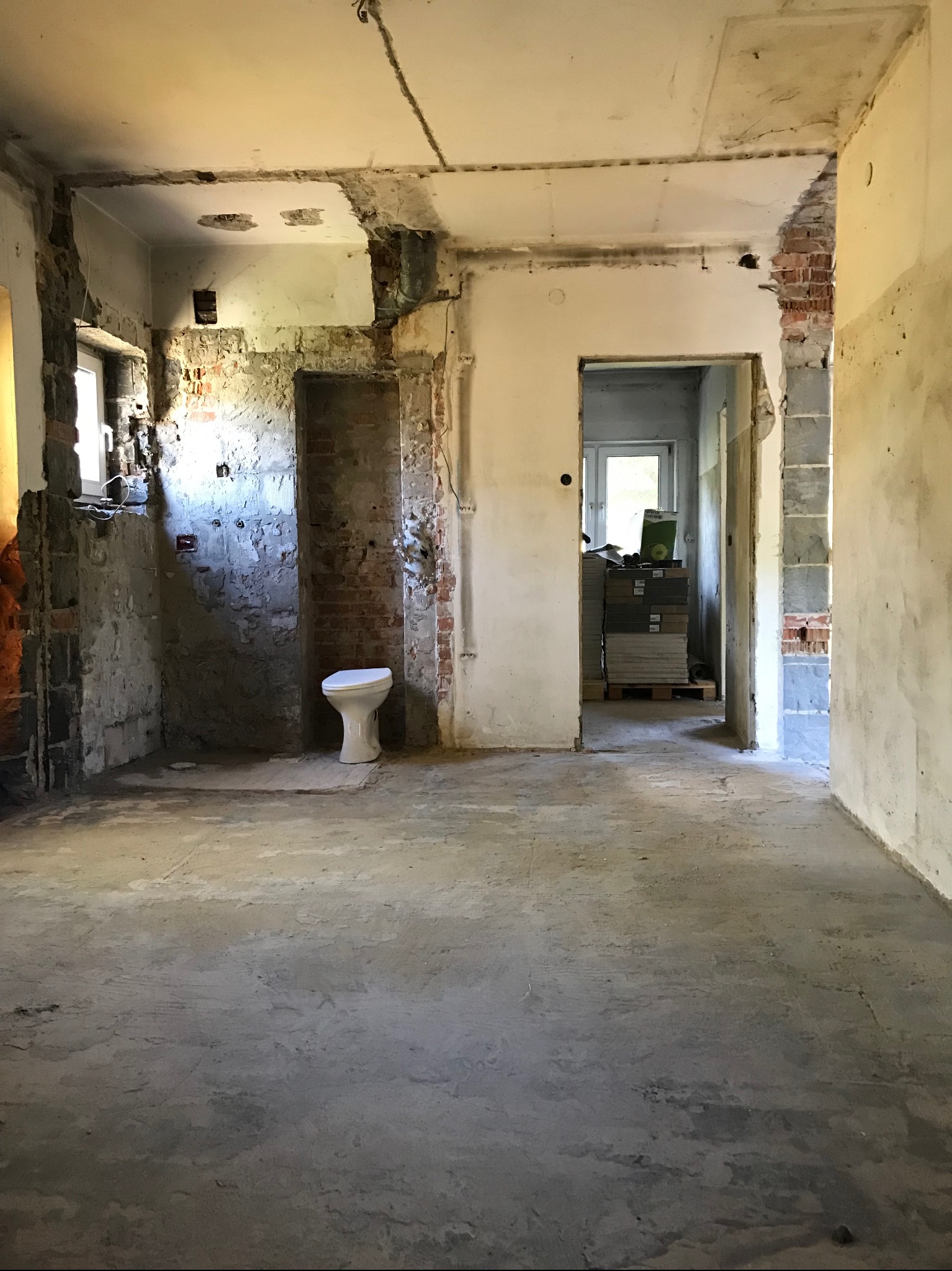 Wnętrze w trakcie remontu: surowe ściany z odsłoniętą cegłą, betonowa podłoga, częściowo usunięte tynki, widoczna toaleta i paleta z płytkami w tle.