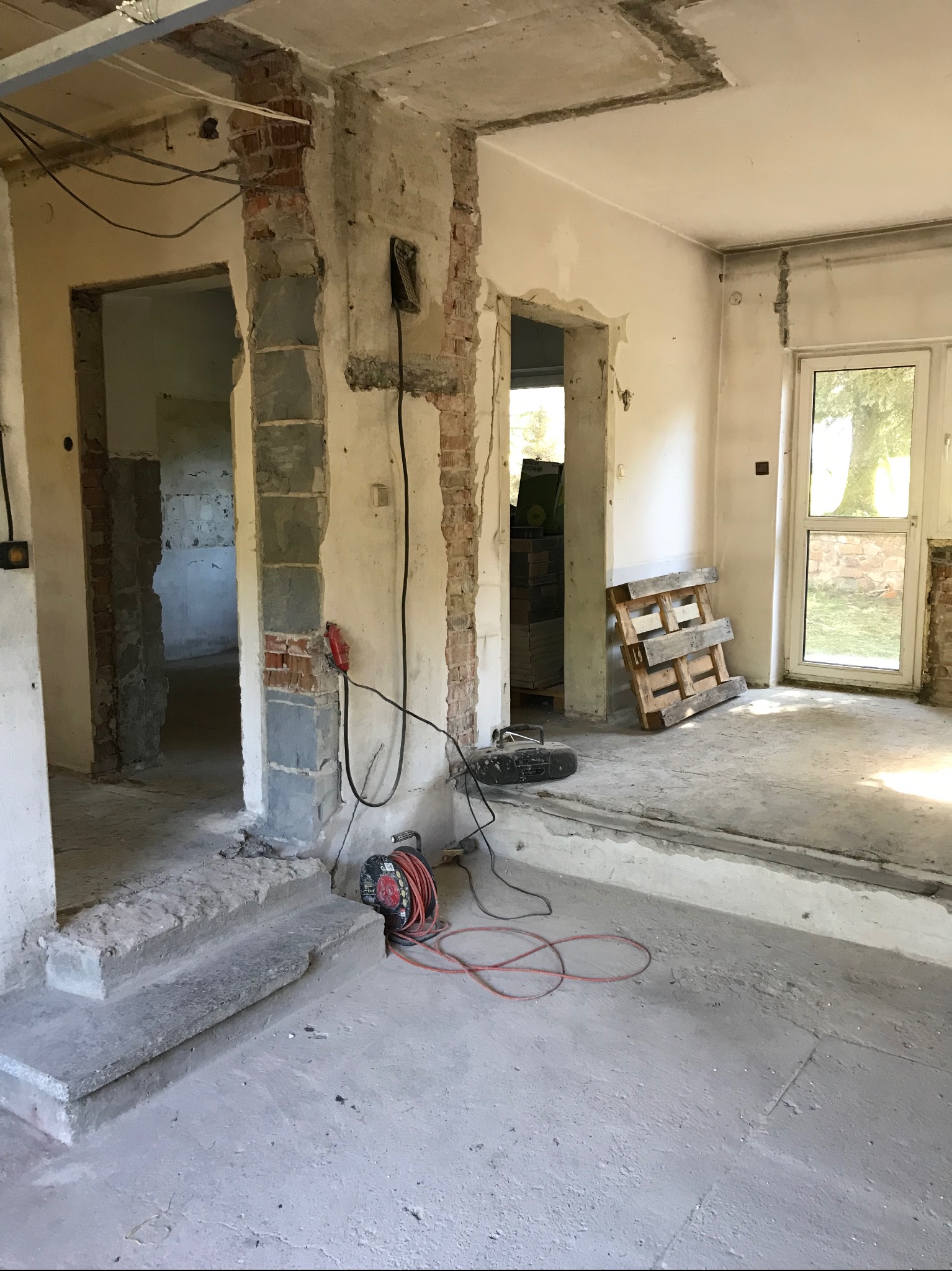 Wnętrze w trakcie gruntownego remontu, widoczne odsłonięte cegły, betonowe schodki wejściowe do pomieszczeń, przewody elektryczne zwisające ze ścian, paleta drewniana oparta o ścianę przy oknie...