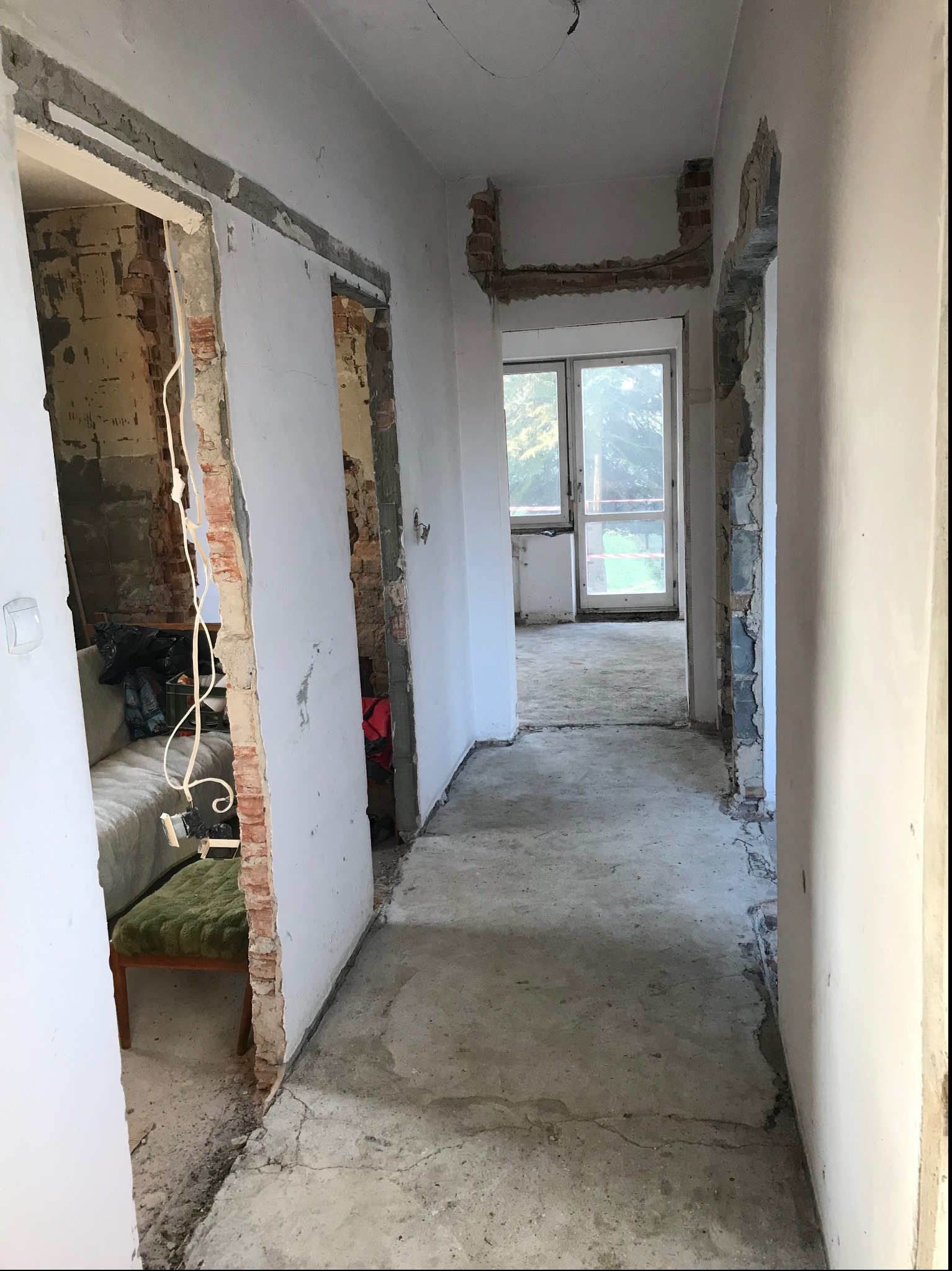 Korytarz w trakcie remontu, z widocznymi fragmentami odsłoniętej cegły wokół otworów drzwiowych, surowy beton na podłodze i przygotowane ściany do dalszych prac wykończeniowych.