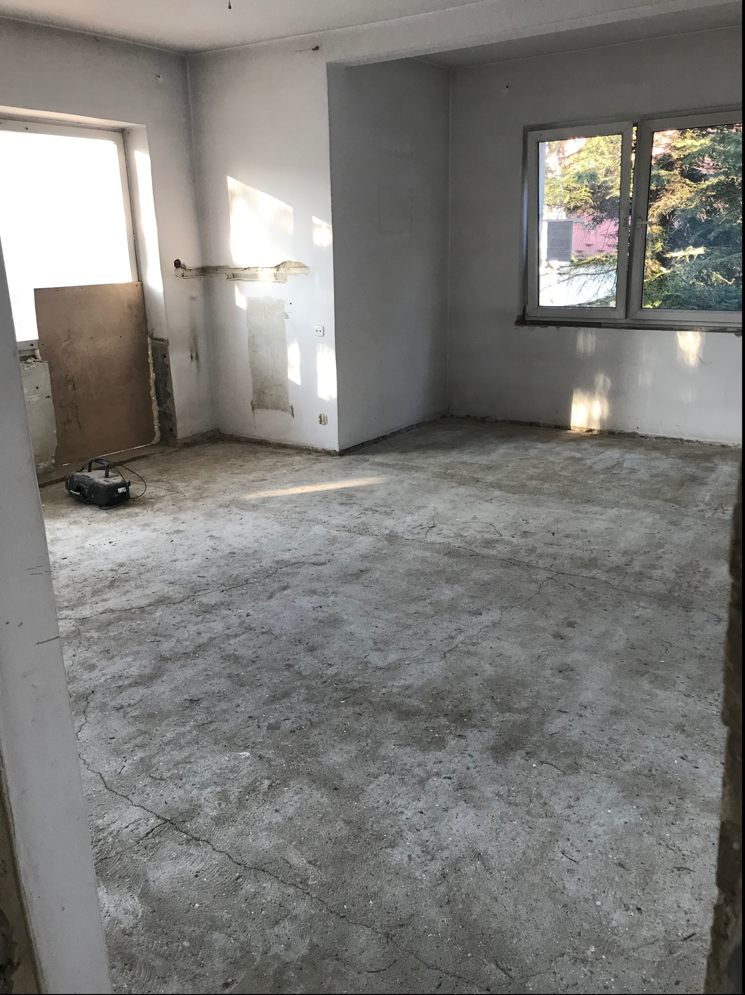 Pokój w trakcie remontu: surowe ściany, betonowa podłoga z pęknięciami, przygotowany do dalszych prac wykończeniowych, widoczne okna i narzędzie pracy.