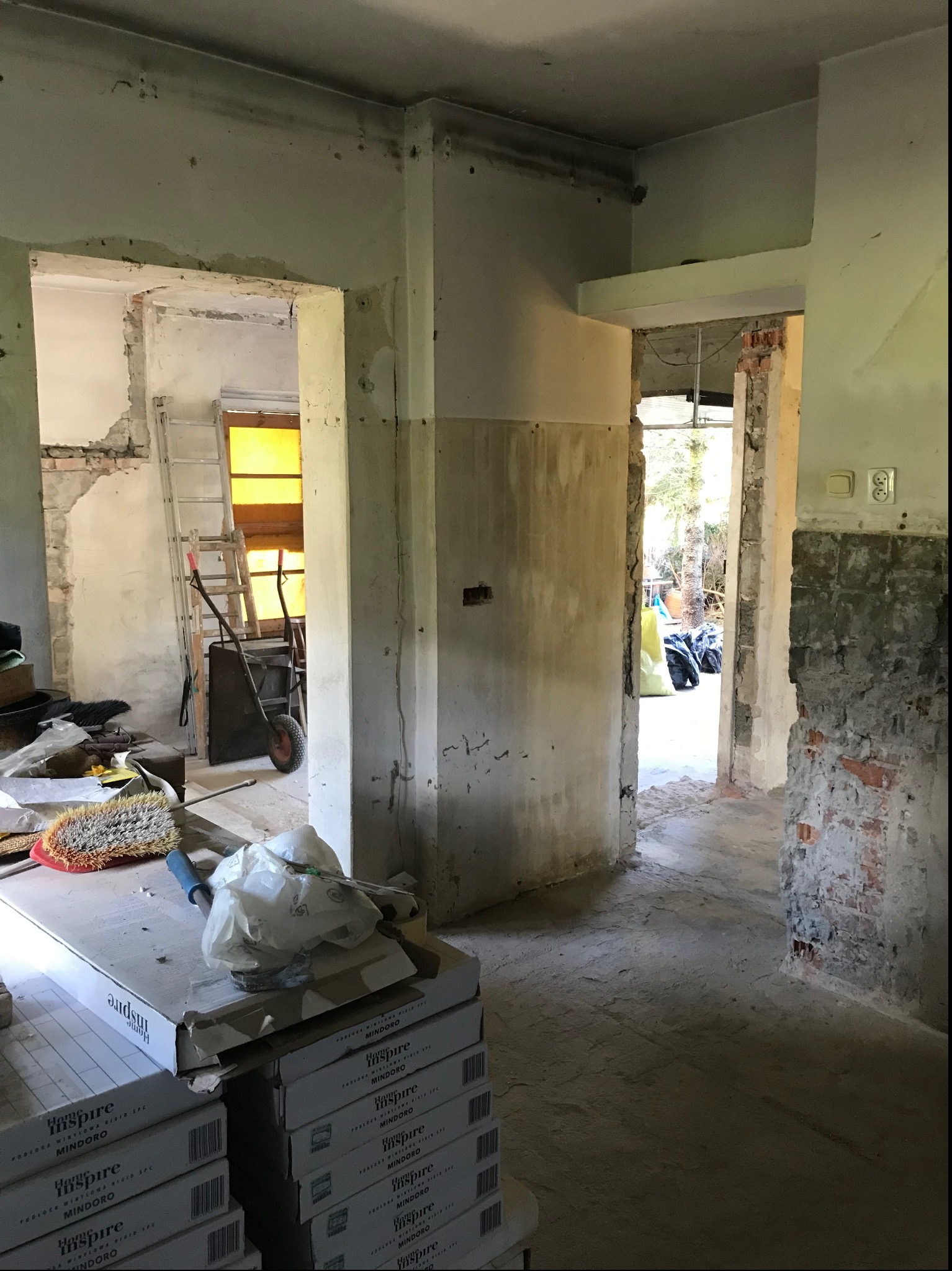Wnętrze w trakcie remontu z widocznymi surowymi ścianami, stertą paneli podłogowych marki Inspire Mindoro, drabiną i wózkiem transportowym w tle.