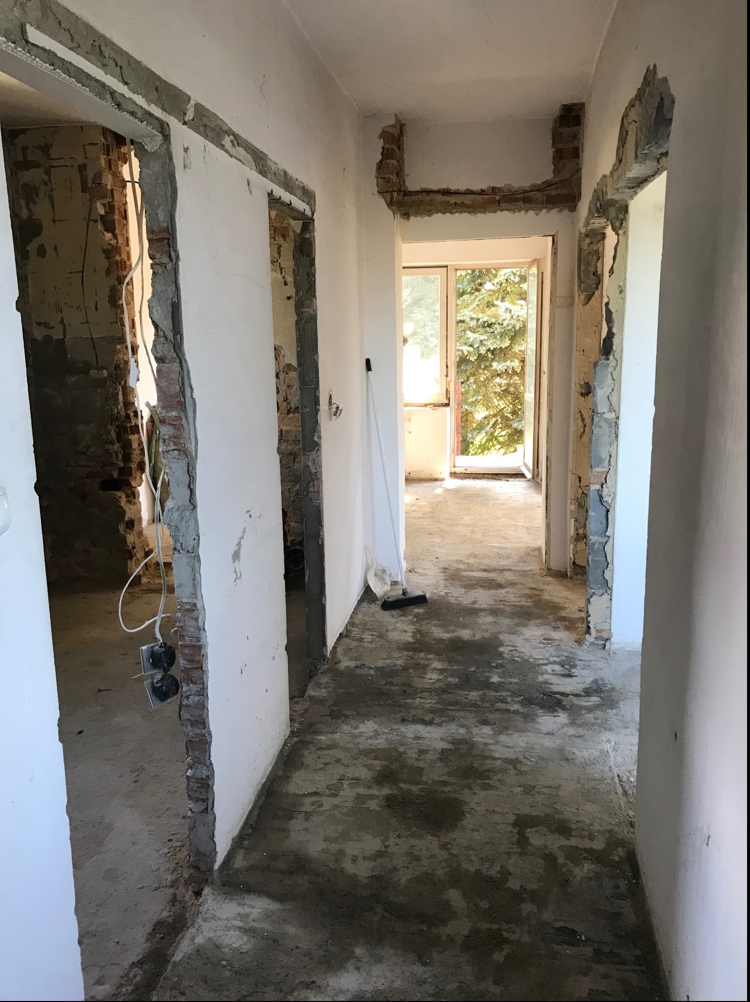 Korytarz w trakcie remontu: surowy beton na podłodze, ściany częściowo otynkowane, widoczne fragmenty cegieł przy ościeżnicach, wystające przewody elektryczne, okno z widokiem na zieleń w oddali.