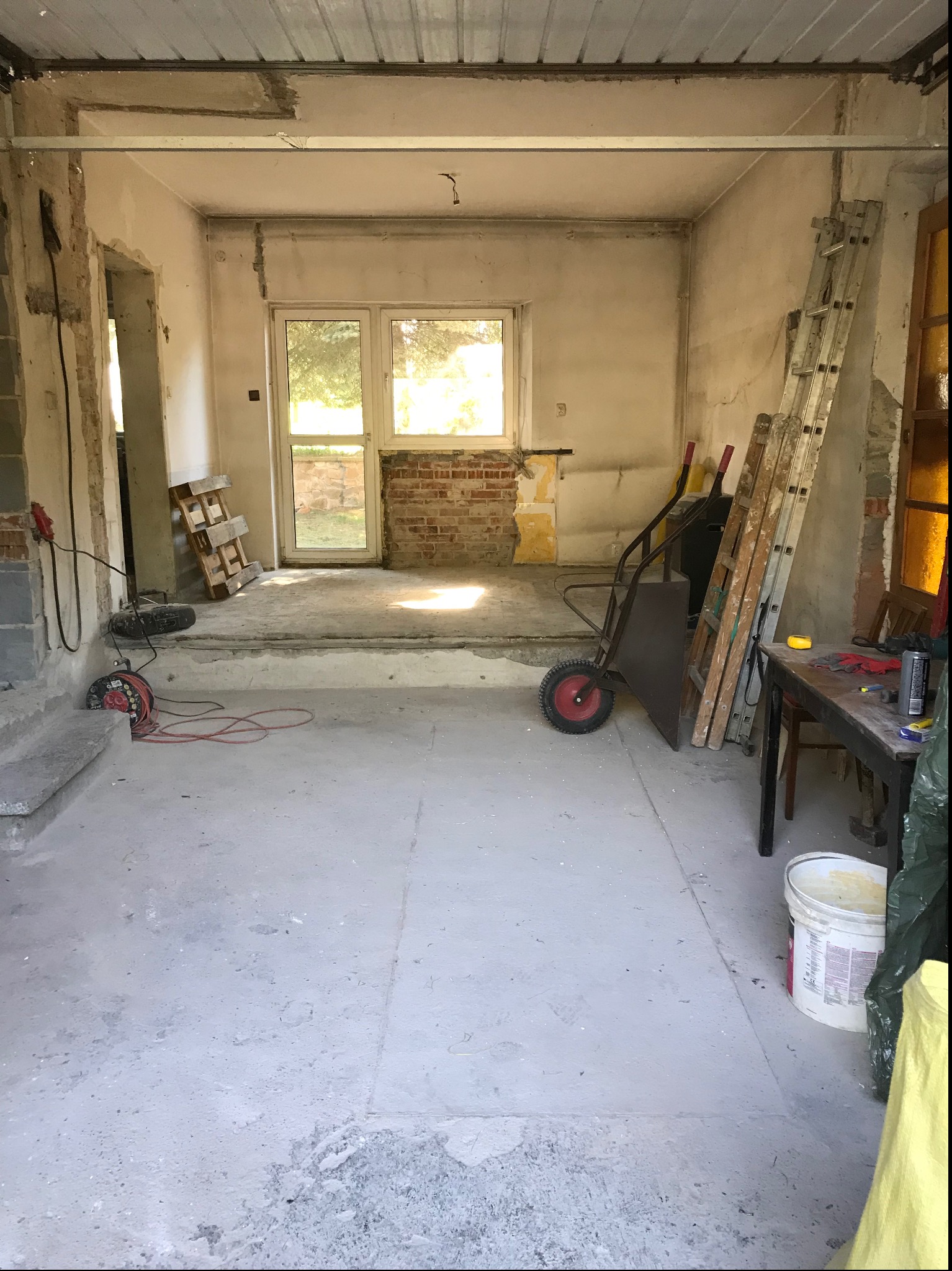 Wnętrze w trakcie remontu: surowe ściany z fragmentami starej farby, odsłonięta cegła przy oknie, prowizoryczne schody, drabina oparta o ścianę, taczka, przedłużacz na podłodze i wiadro farby...