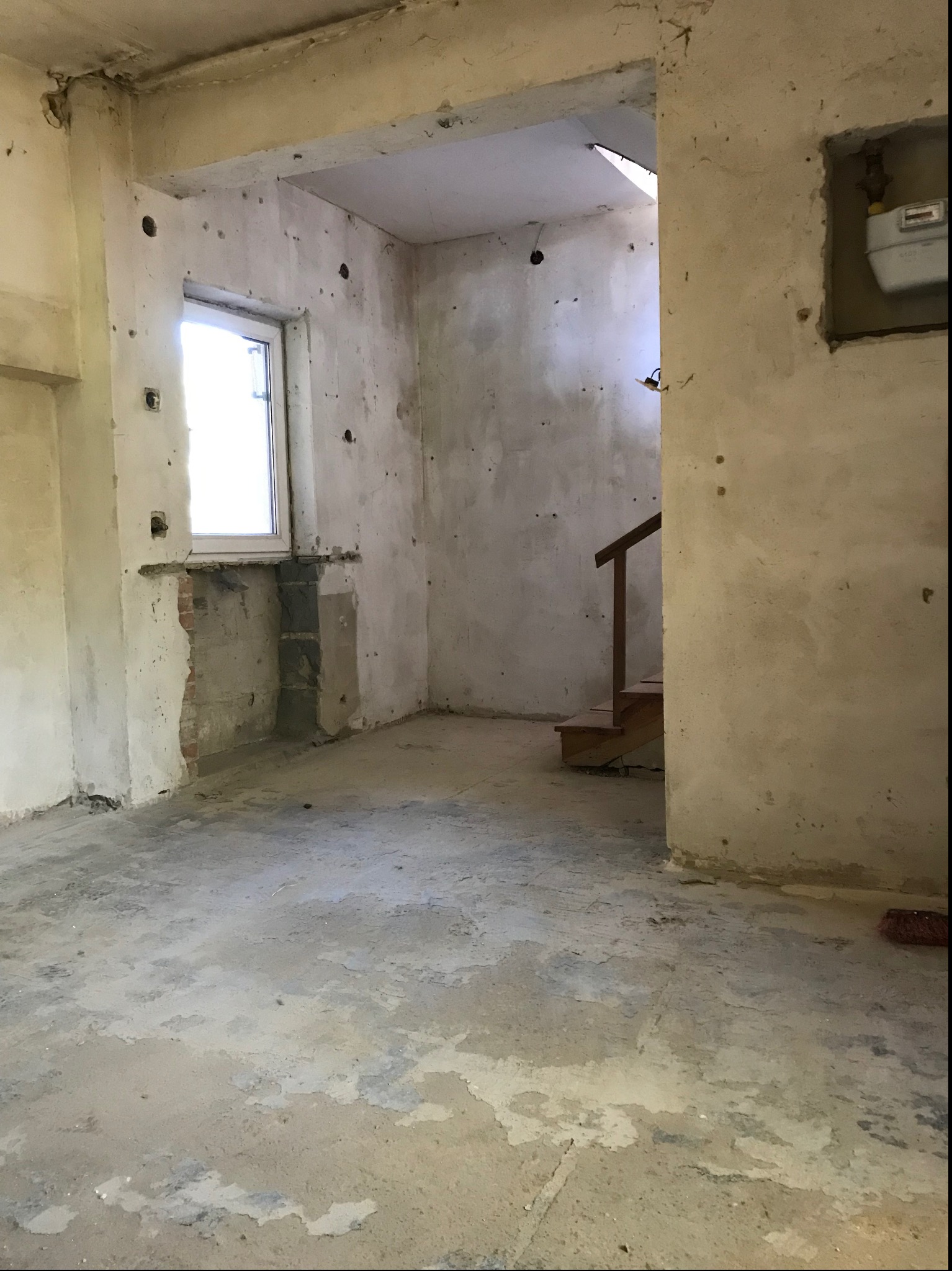 Wnętrze w trakcie prac przygotowawczych do wykończenia: surowe ściany z widocznymi otworami, fragmenty odsłoniętej cegły pod oknem, betonowa podłoga z plamami, widoczne schody w głębi.