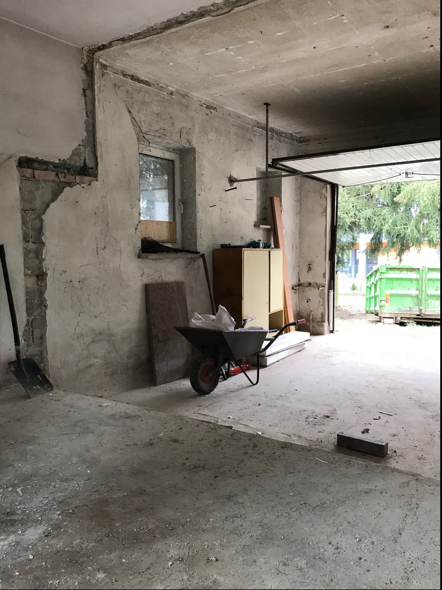 Wnętrze remontowanego pomieszczenia z odsłoniętą ścianą z cegieł, taczką wypełnioną materiałami budowlanymi i otwartą bramą garażową