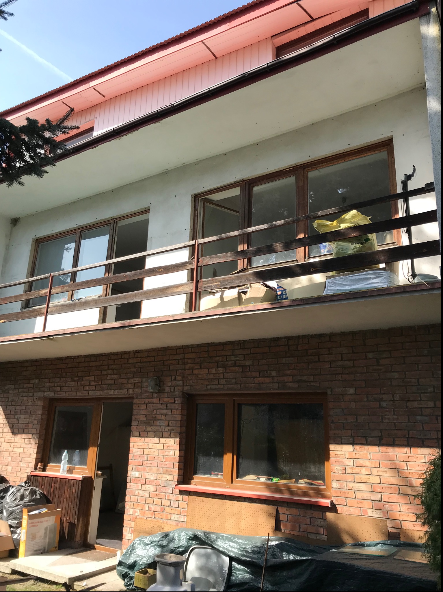 Dwupiętrowy dom z ceglaną elewacją na parterze i ocieplonym poddaszem, z drewnianą balustradą balkonu i widocznymi materiałami budowlanymi