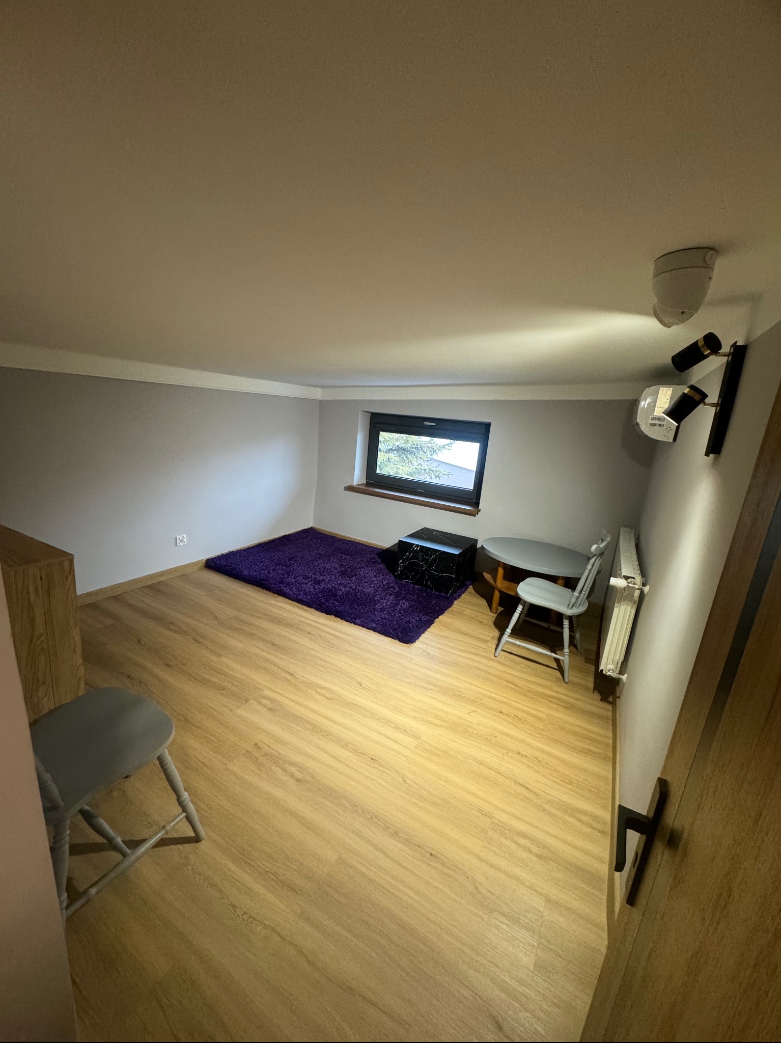 Wnętrze pokoju z jasnymi panelami podłogowymi, szarą ścianą, fioletowym dywanem, małym oknem, stolikiem i krzesłem w stylu vintage oraz nowoczesnym oświetleniem ściennym.