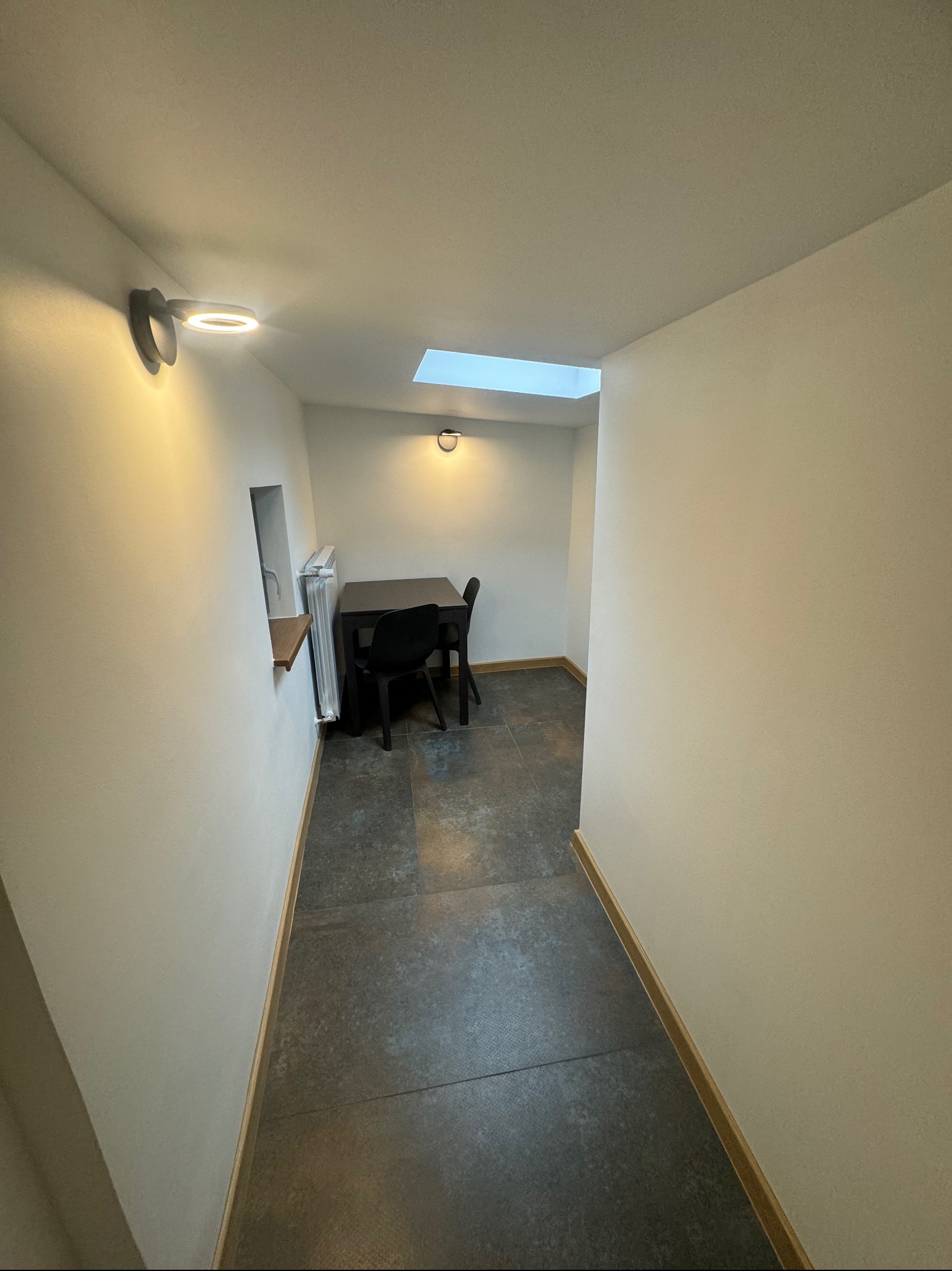 Wnętrze korytarza z szarymi płytkami podłogowymi, białymi ścianami oświetlonymi lampami punktowymi, z widokiem na mały pokój z oknem dachowym, czarnym stołem i krzesłami oraz grzejnikiem.