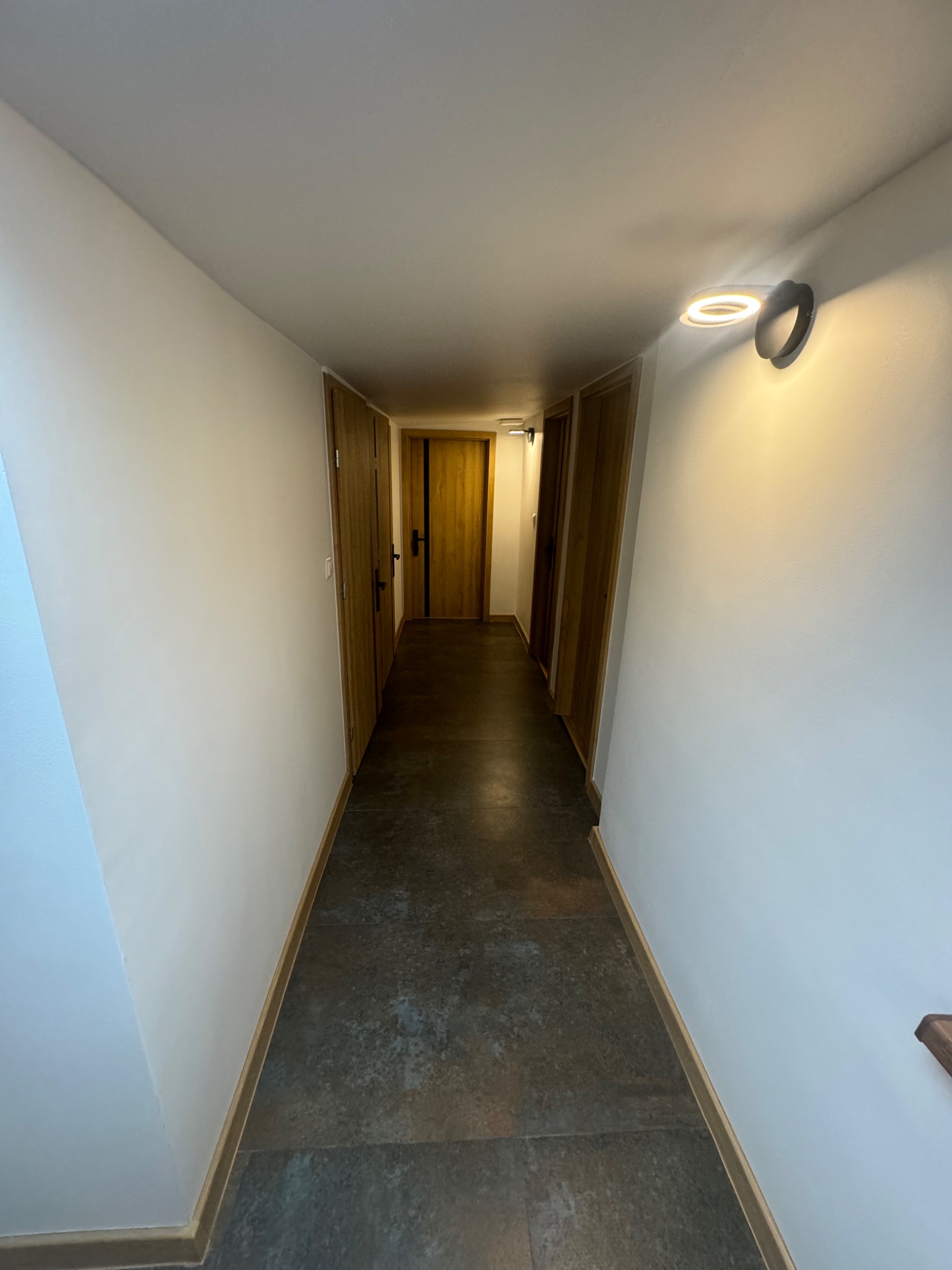 Wnętrze korytarza z białymi ścianami, szarą podłogą z płytek imitujących kamień, drewnianymi drzwiami i nowoczesnym okrągłym oświetleniem ściennym.