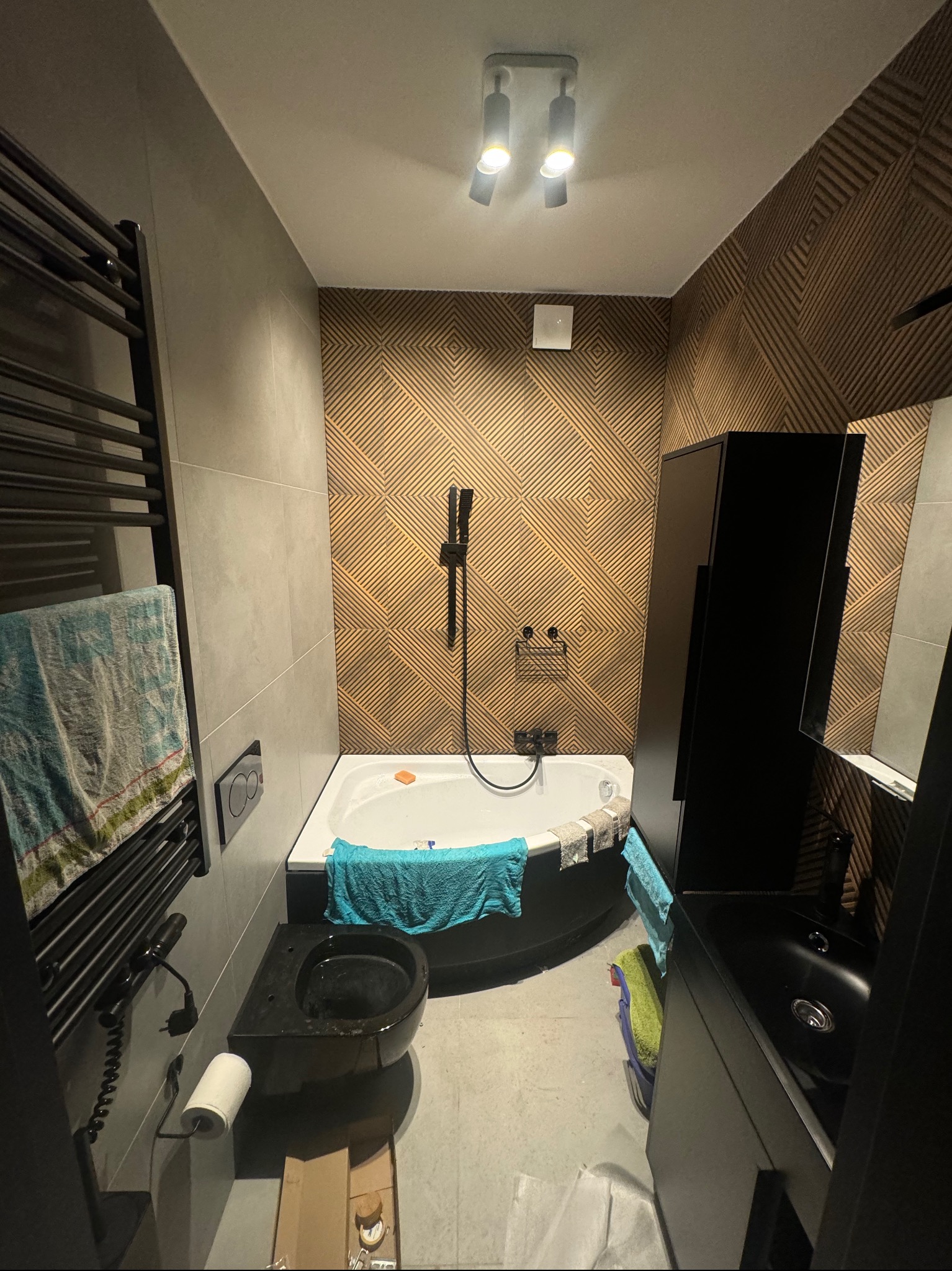 Nowoczesna łazienka z geometryczną ścianą, czarną armaturą i grzejnikiem drabinkowym, widok na wannę z hydromasażem i wiszącą toaletę.