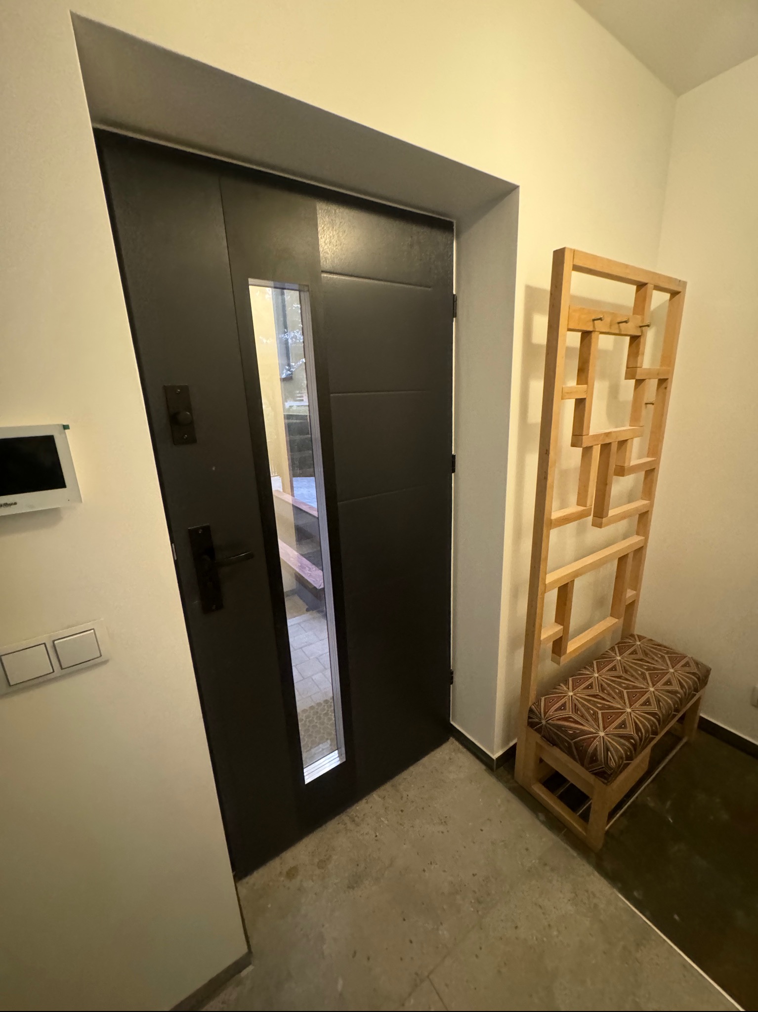 Nowoczesny przedpokój z ciemnymi drzwiami wejściowymi z przeszkleniem i drewnianym wieszakiem z siedziskiem w geometryczne wzory.