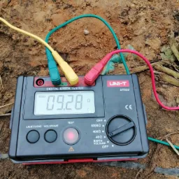 Miernik rezystancji uziemienia UNI-T UT522 z podłączonymi przewodami pomiarowymi wbitymi w ziemię, wyświetlający wynik 09.28 omów, w tle widoczna gleba.