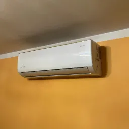 Zamontowany na pomarańczowej ścianie, biały klimatyzator Rotenso RONI Series z delikatnym złotym akcentem na górnej krawędzi.