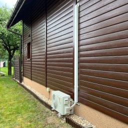 KLIMERA Michał Garbiec - Boczny widok brązowego, drewnianego domu z zamontowaną białą jednostką zewnętrzną klimatyzacji, rurą odprowadzającą i ozdobnym obramowaniem z kamieni i palisady.