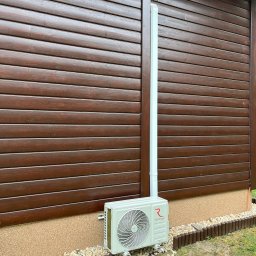 KLIMERA Michał Garbiec - Zamontowana jednostka zewnętrzna klimatyzacji Rotenso na brązowej elewacji drewnianej, z widoczną białą rurą odprowadzającą. Estetyczne wykończenie rabaty.