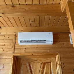 KLIMERA Michał Garbiec - Biały klimatyzator Rotenso zamontowany na drewnianej ścianie nad drzwiami w pomieszczeniu wykończonym boazerią. Widok z dołu.