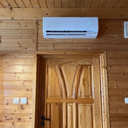 KLIMERA Michał Garbiec - Biały klimatyzator Roni zamontowany nad drewnianymi drzwiami w pomieszczeniu wykończonym boazerią. Widoczne włączniki światła po lewej stronie.