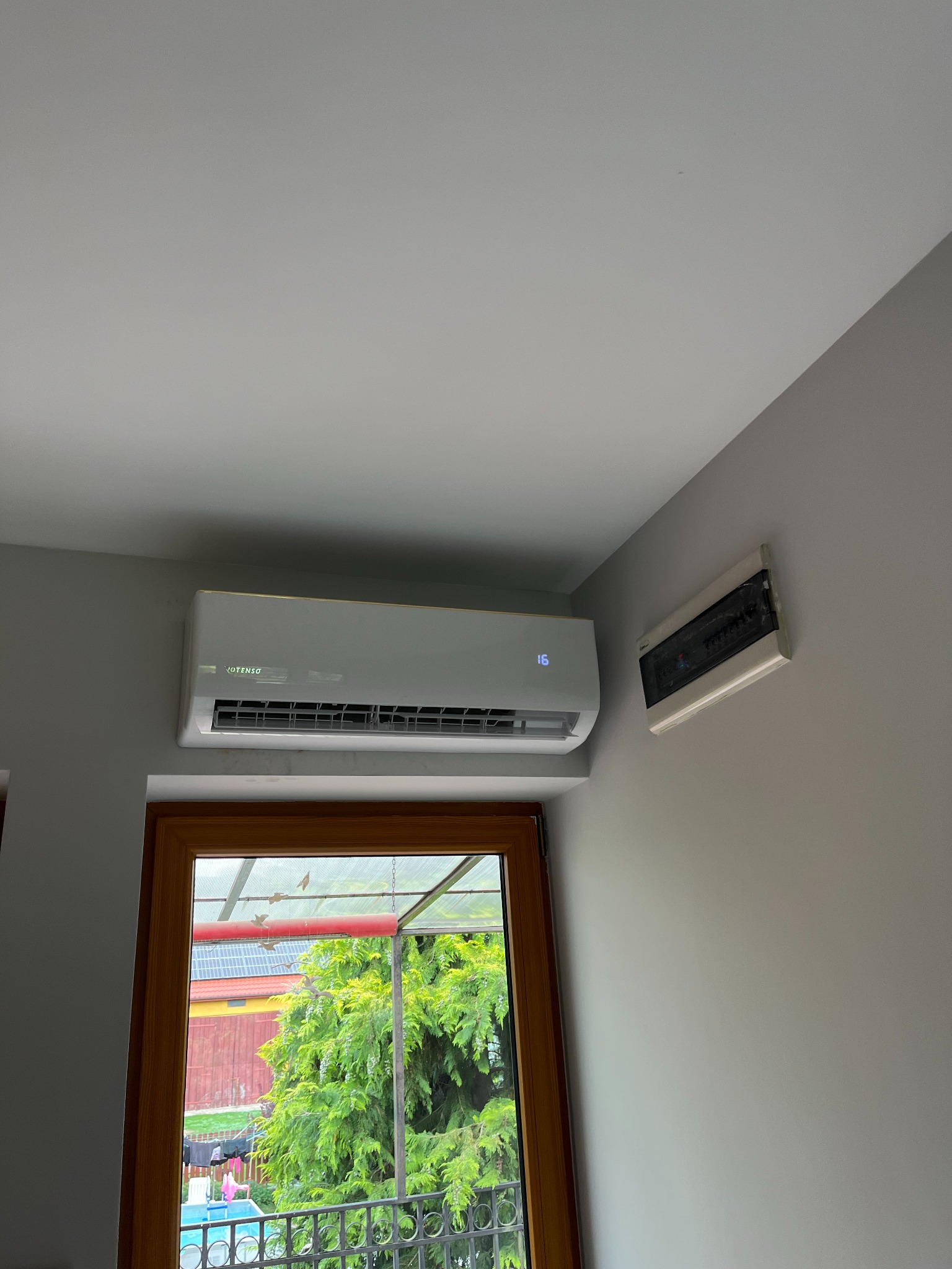 Biały klimatyzator marki VOTENSO zamontowany na szarej ścianie obok skrzynki elektrycznej, widok z okna na zewnątrz.