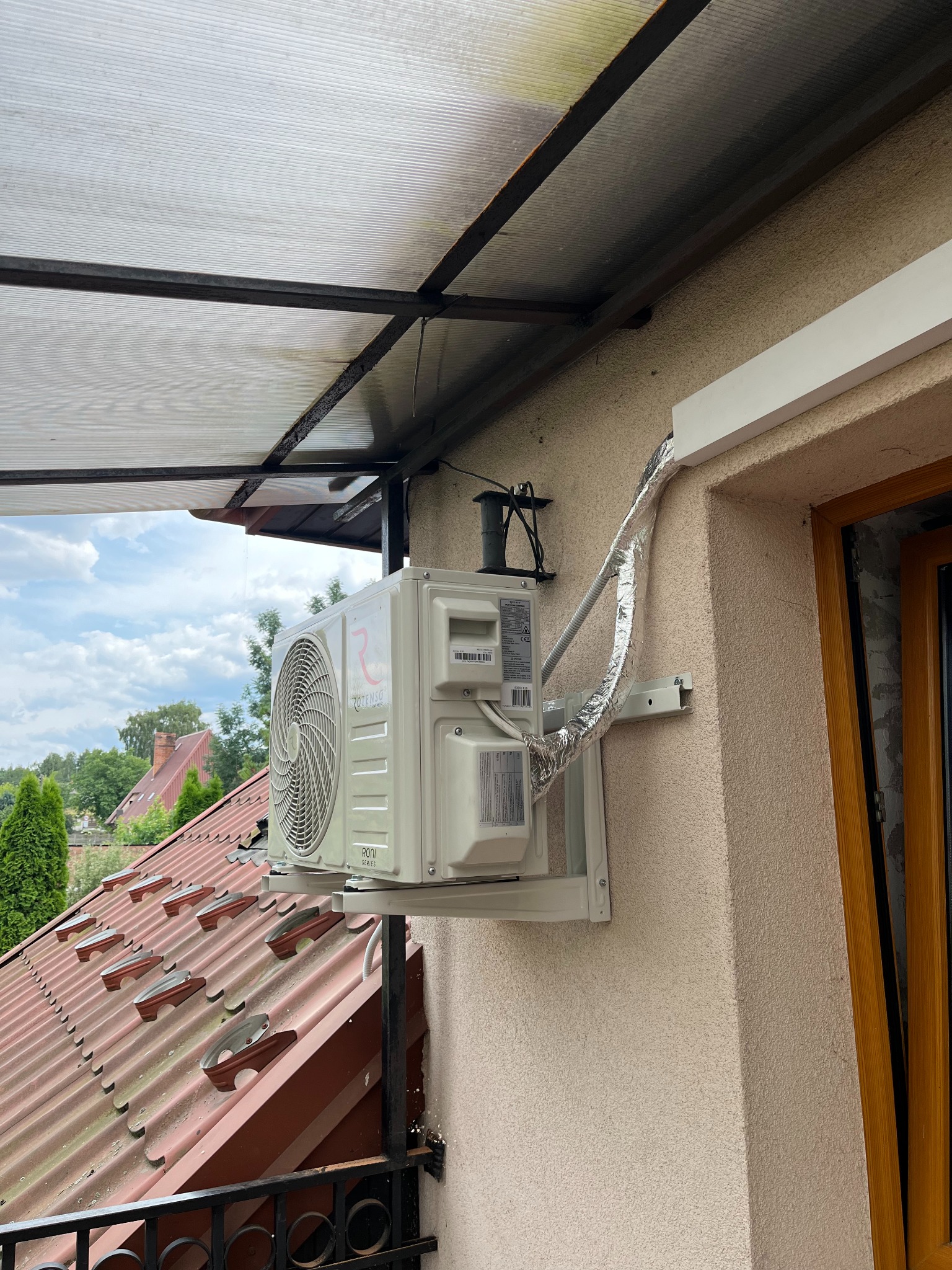Zamontowana jednostka zewnętrzna klimatyzacji Rotenso Roni Series na elewacji budynku z widocznymi przewodami w izolacji, balkonem i dachem w tle.