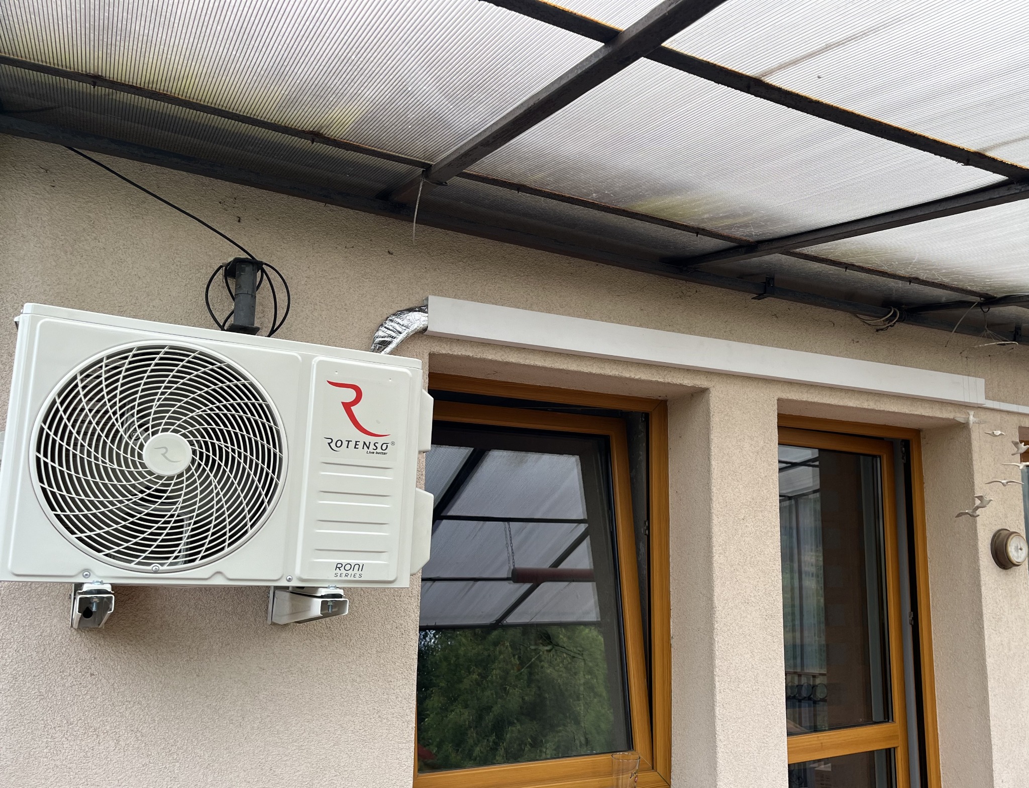 Biała jednostka zewnętrzna klimatyzacji Rotenso Roni Series zamontowana na ścianie budynku pod zadaszeniem z poliwęglanu, widoczne okno z drewnianą ramą i elementy dekoracyjne na elewacji.