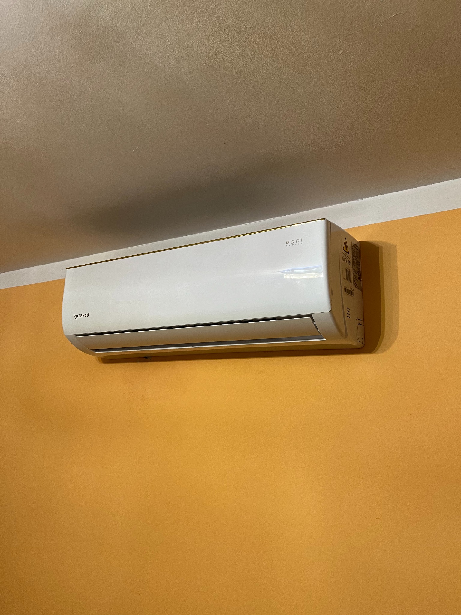 Zamontowany na pomarańczowej ścianie, biały klimatyzator Rotenso RONI Series z delikatnym złotym akcentem na górnej krawędzi.