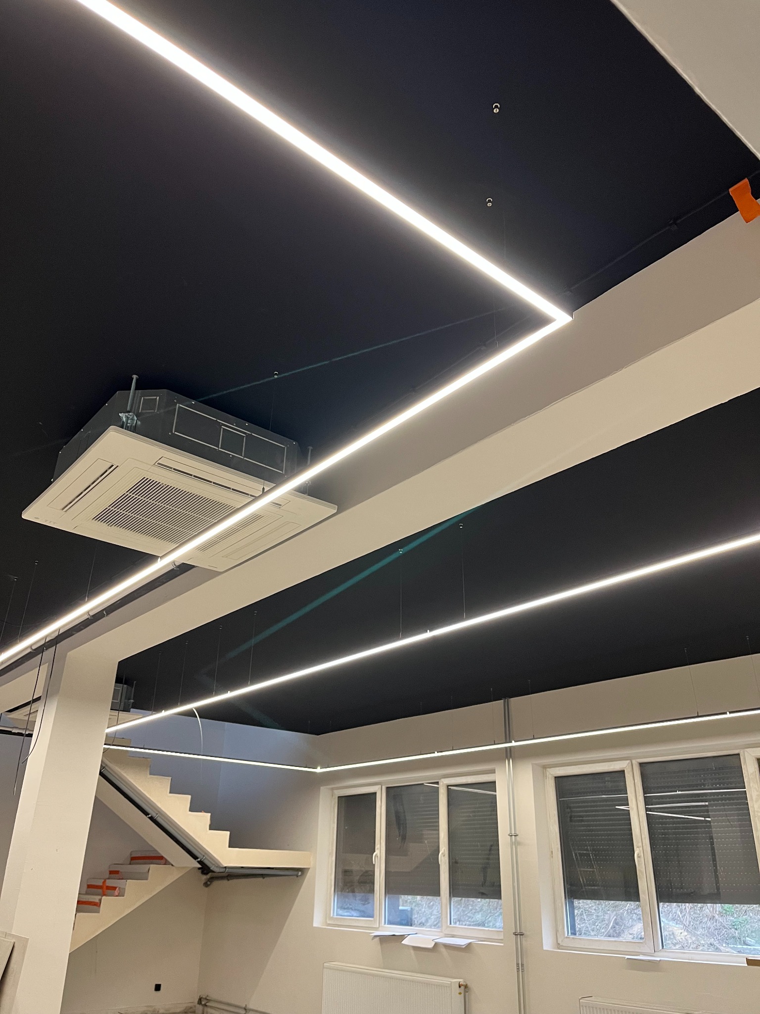 Nowoczesne oświetlenie LED w biurze z czarnym sufitem i klimatyzacją kasetonową. Widoczne schody i okna z żaluzjami. Minimalistyczny design.