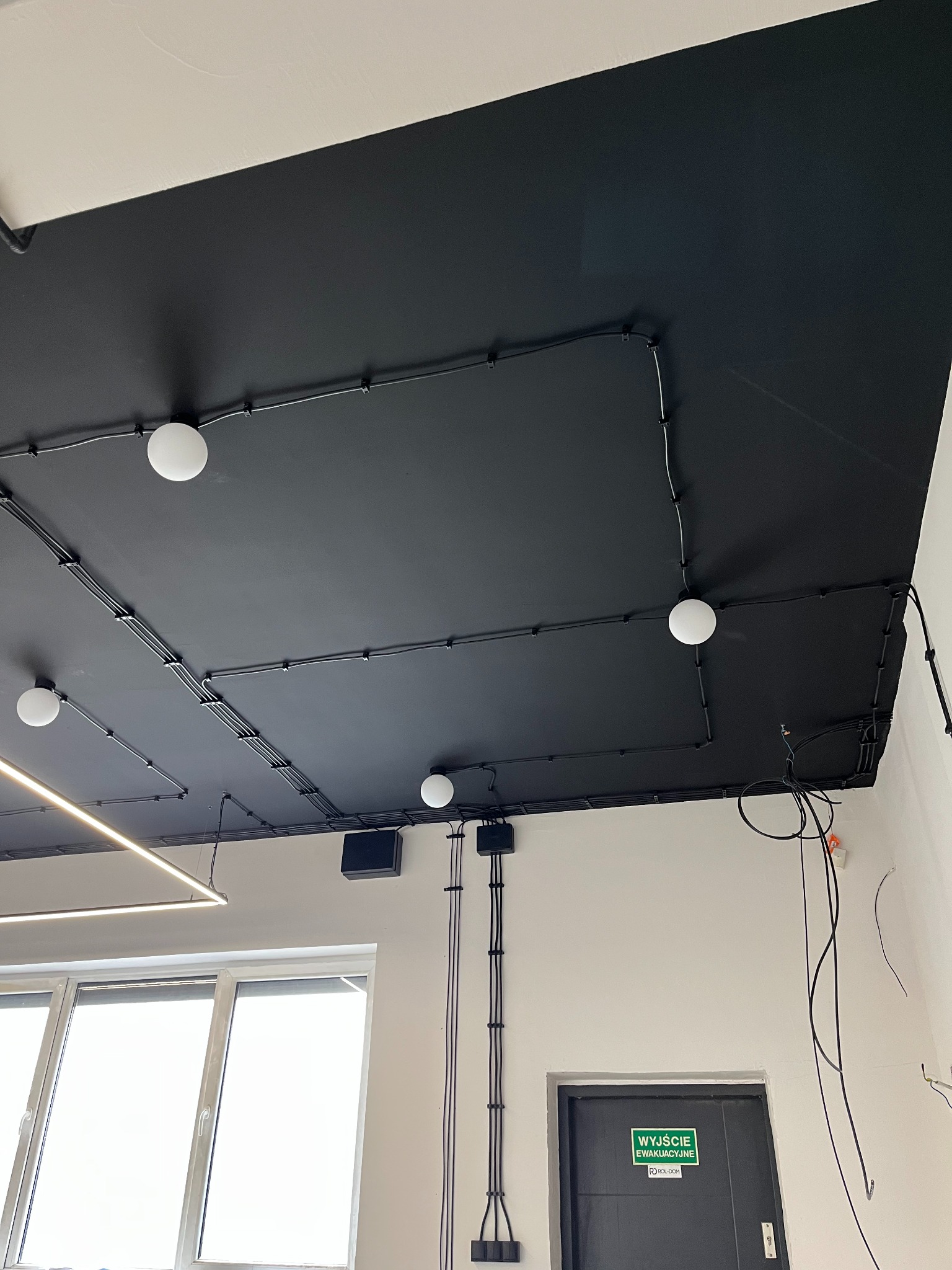 Czarny sufit z widocznymi kablami elektrycznymi prowadzonymi natynkowo, zamocowanymi uchwytami. Widoczne lampy kuliste i liniowe. Instalacja w pomieszczeniu biurowym.