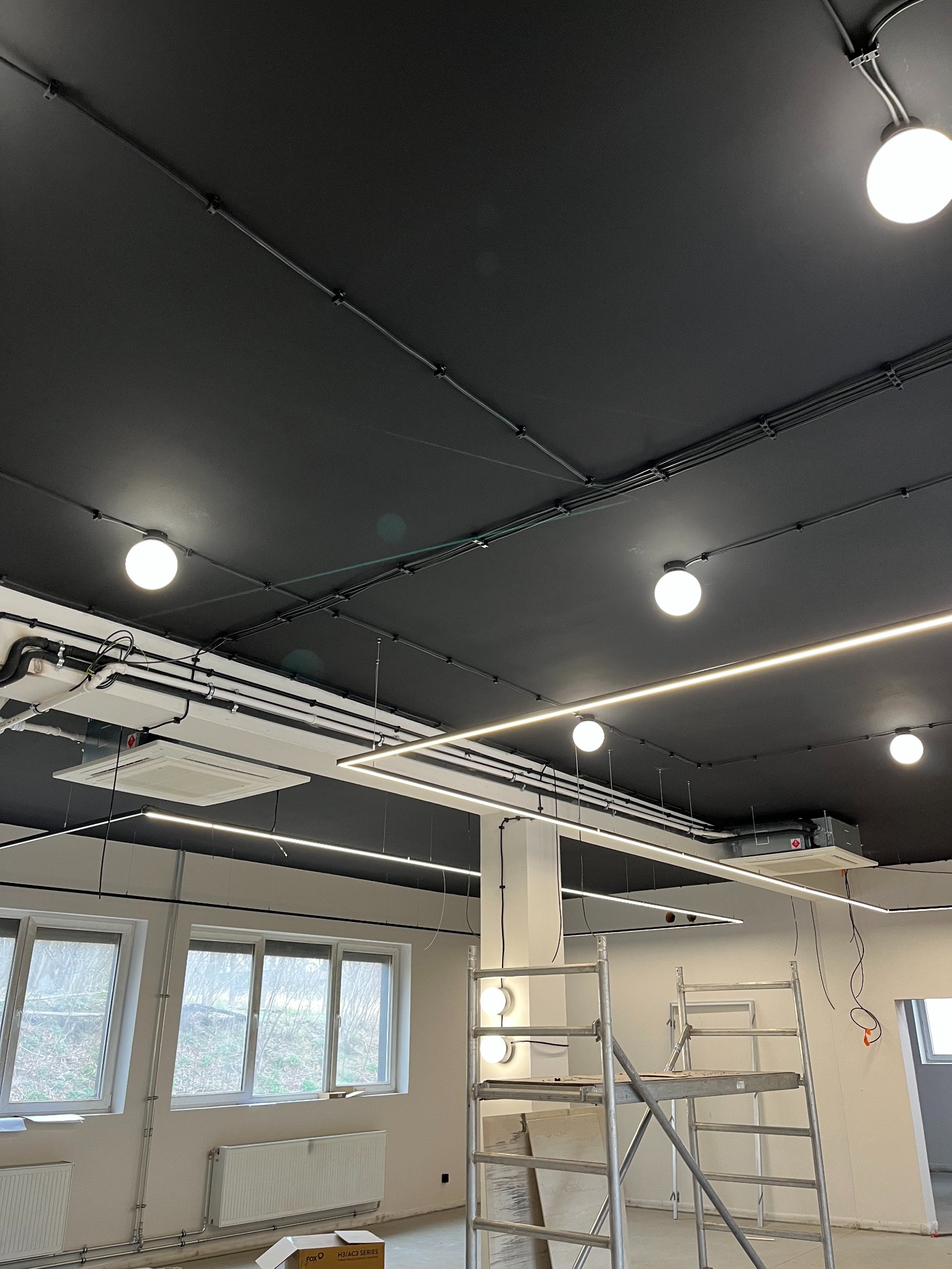 Czarne sufity z widocznymi instalacjami: elektrycznymi, klimatyzacyjnymi i oświetleniowymi. Na pierwszym planie rusztowanie, okna i grzejniki. W tle widoczne drzewa.