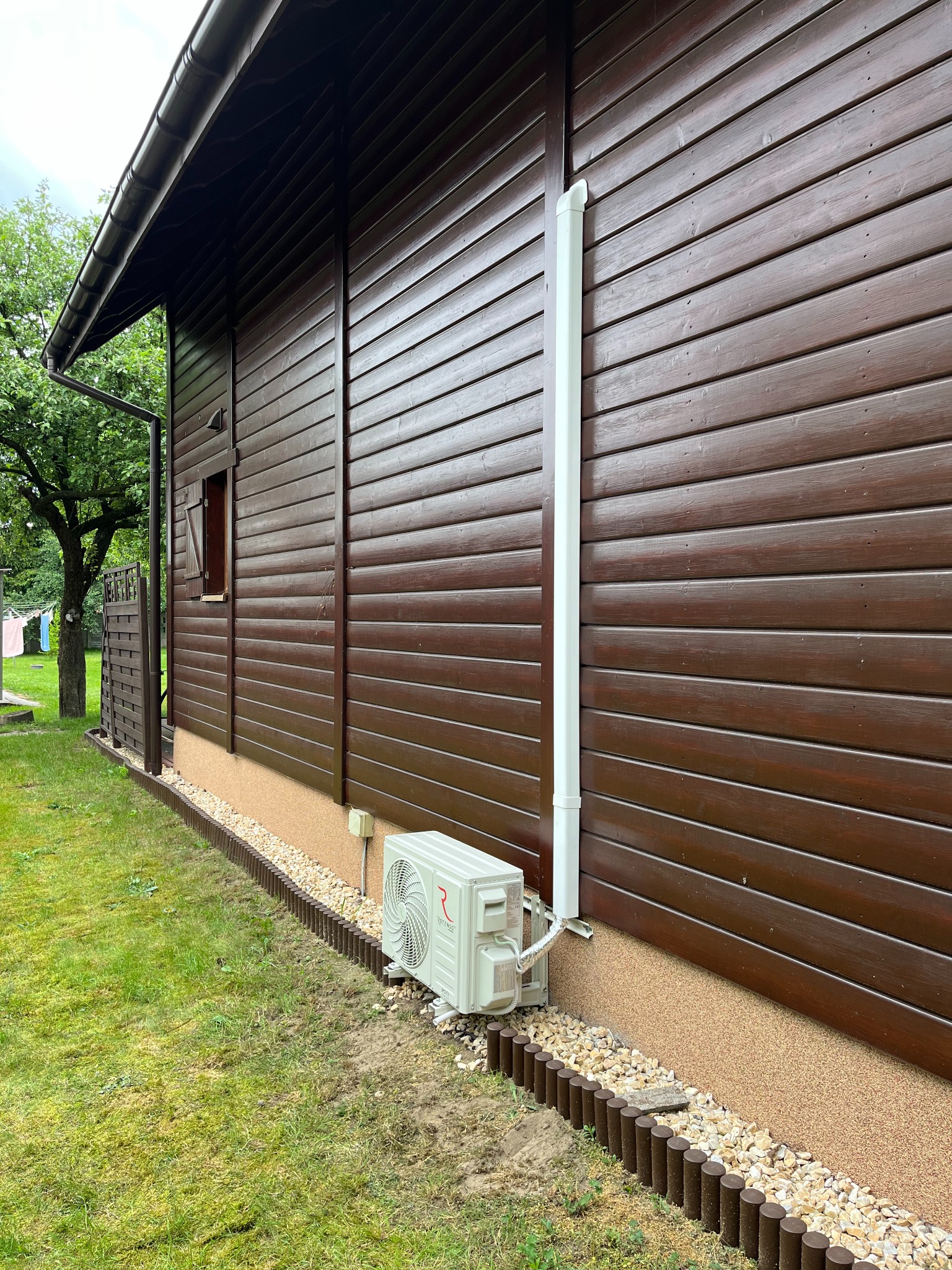 Boczny widok brązowego, drewnianego domu z zamontowaną białą jednostką zewnętrzną klimatyzacji, rurą odprowadzającą i ozdobnym obramowaniem z kamieni i palisady.