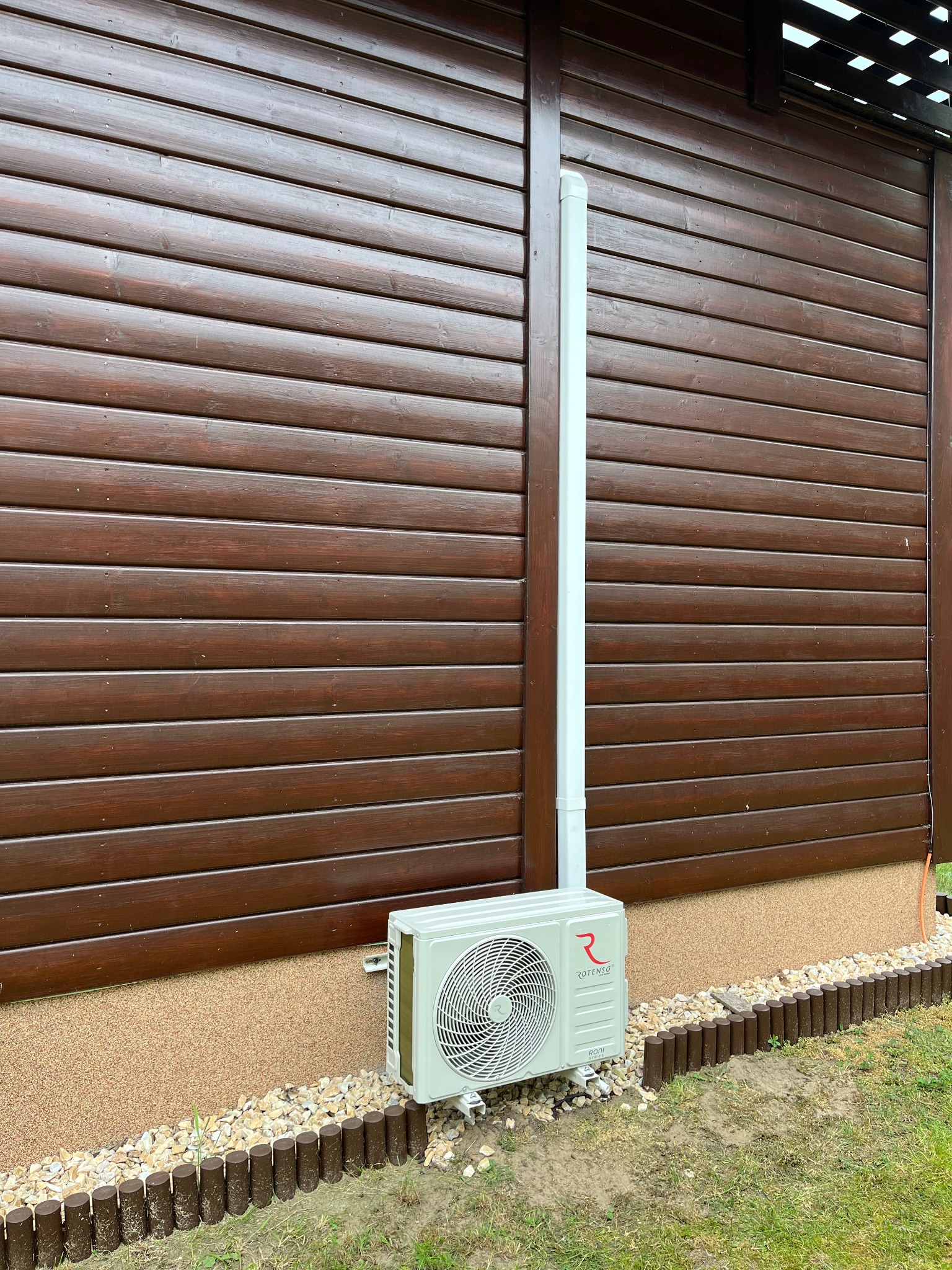 Zamontowana jednostka zewnętrzna klimatyzacji Rotenso na brązowej elewacji drewnianej, z widoczną białą rurą odprowadzającą. Estetyczne wykończenie rabaty.
