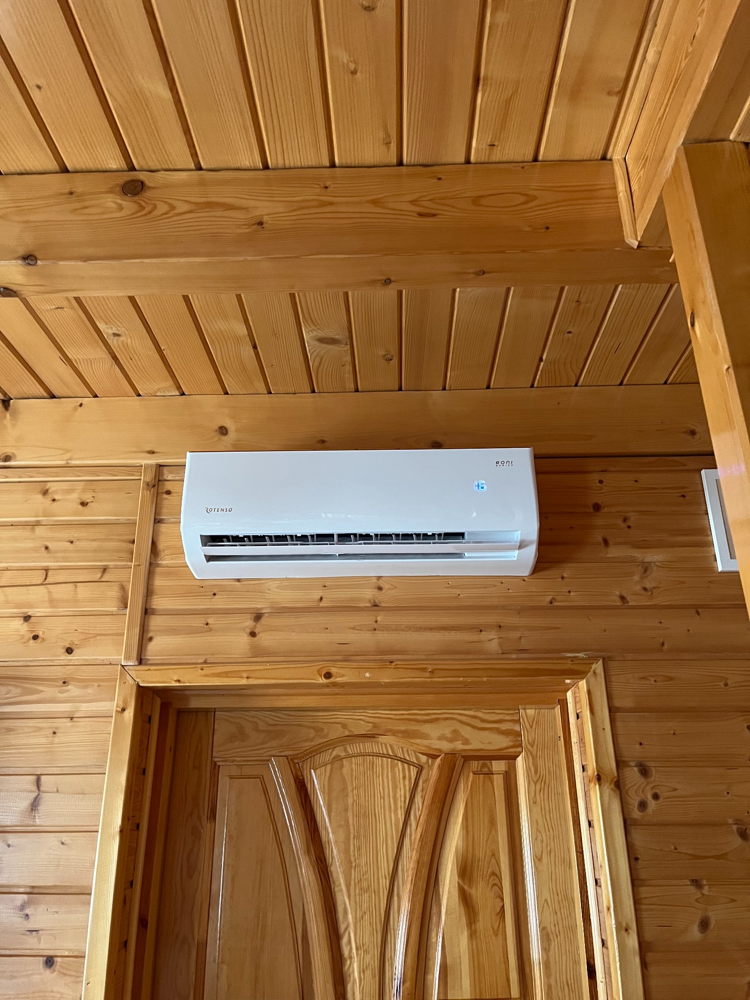 Biały klimatyzator Rotenso zamontowany na drewnianej ścianie nad drzwiami w pomieszczeniu wykończonym boazerią. Widok z dołu.