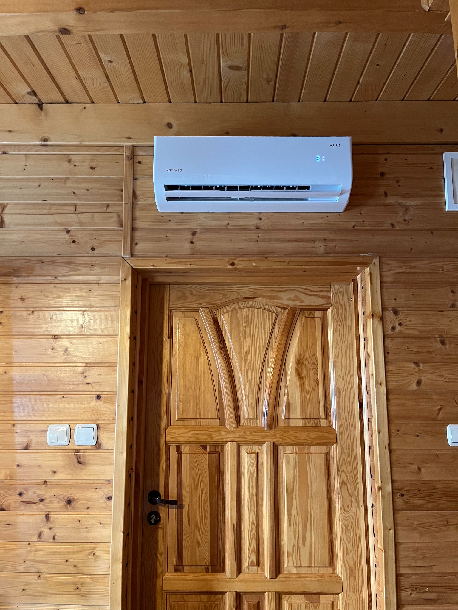 Biały klimatyzator Roni zamontowany nad drewnianymi drzwiami w pomieszczeniu wykończonym boazerią. Widoczne włączniki światła po lewej stronie.