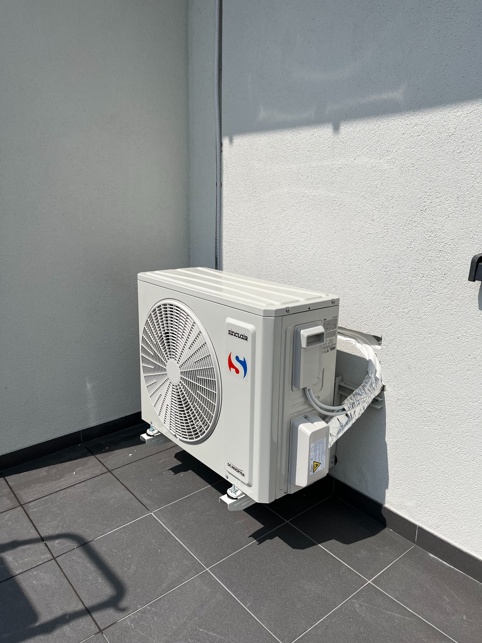 Biała jednostka zewnętrzna klimatyzacji Sinclair zamontowana na szarym balkonie, z widocznymi przewodami w srebrnej izolacji i fragmentem rury odprowadzającej skropliny.