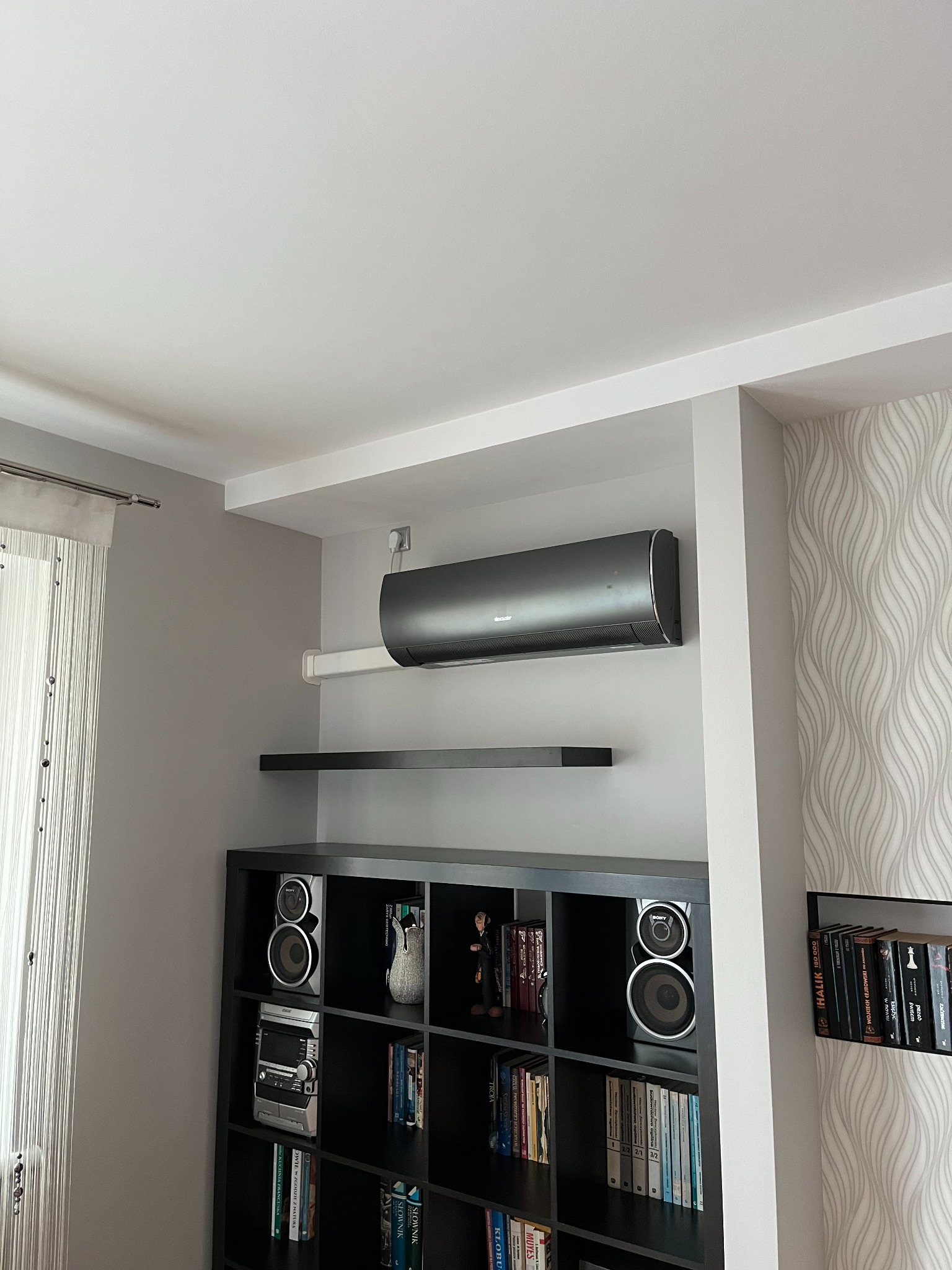 Czarny klimatyzator ścienny marki Kaisai zamontowany nad czarnym regałem z książkami i sprzętem audio w nowoczesnym wnętrzu z wzorzystą tapetą.