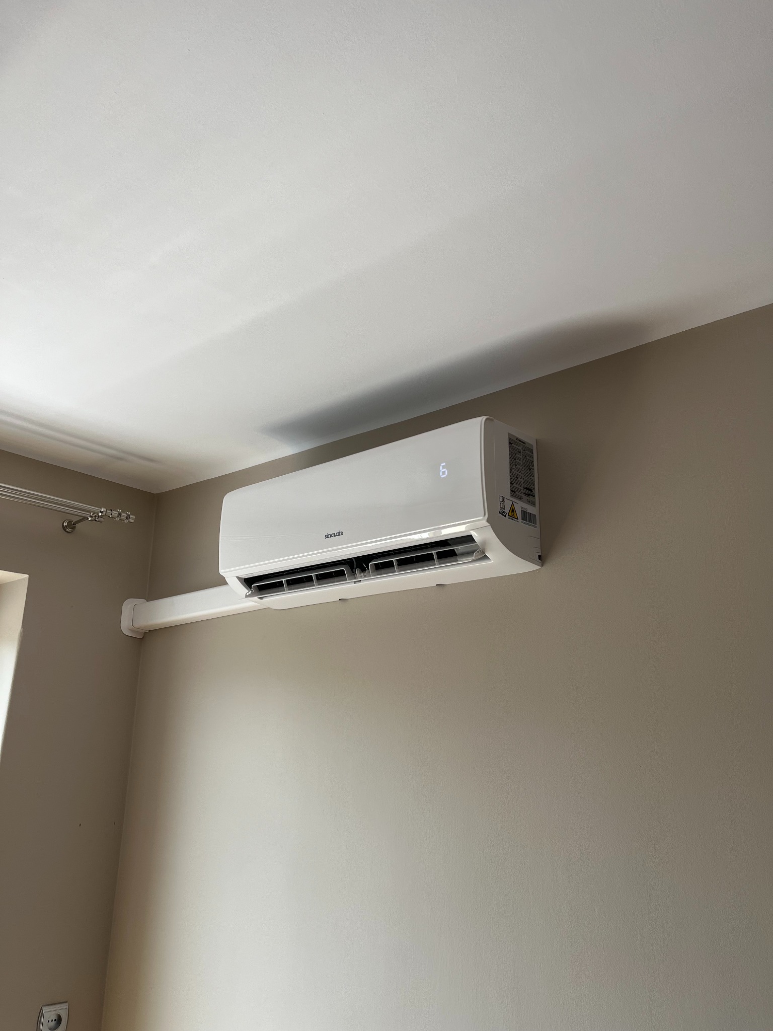 Biały klimatyzator Sinclair zamontowany na beżowej ścianie w pokoju, widoczny fragment karnisza przy oknie. Urządzenie z wyświetlaczem cyfrowym.