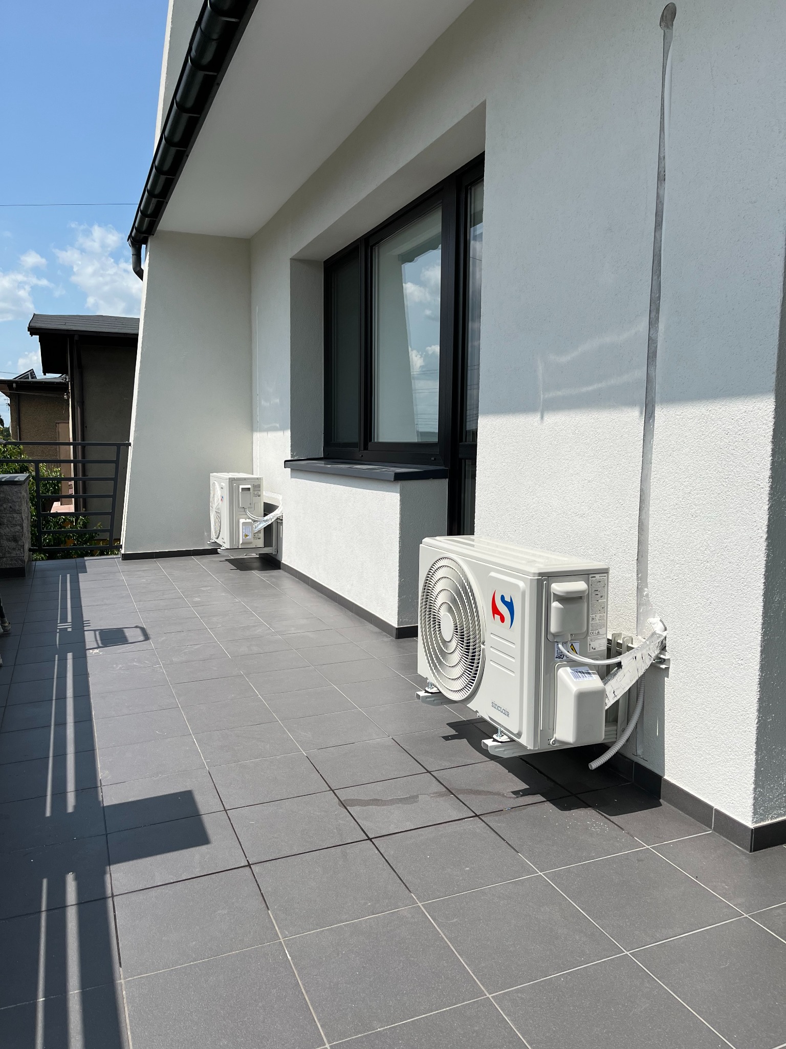 Biała elewacja budynku z dwoma zamontowanymi klimatyzatorami Sinclair na balkonie z szarymi płytkami. Widoczne okno i rynna. Słoneczny dzień.