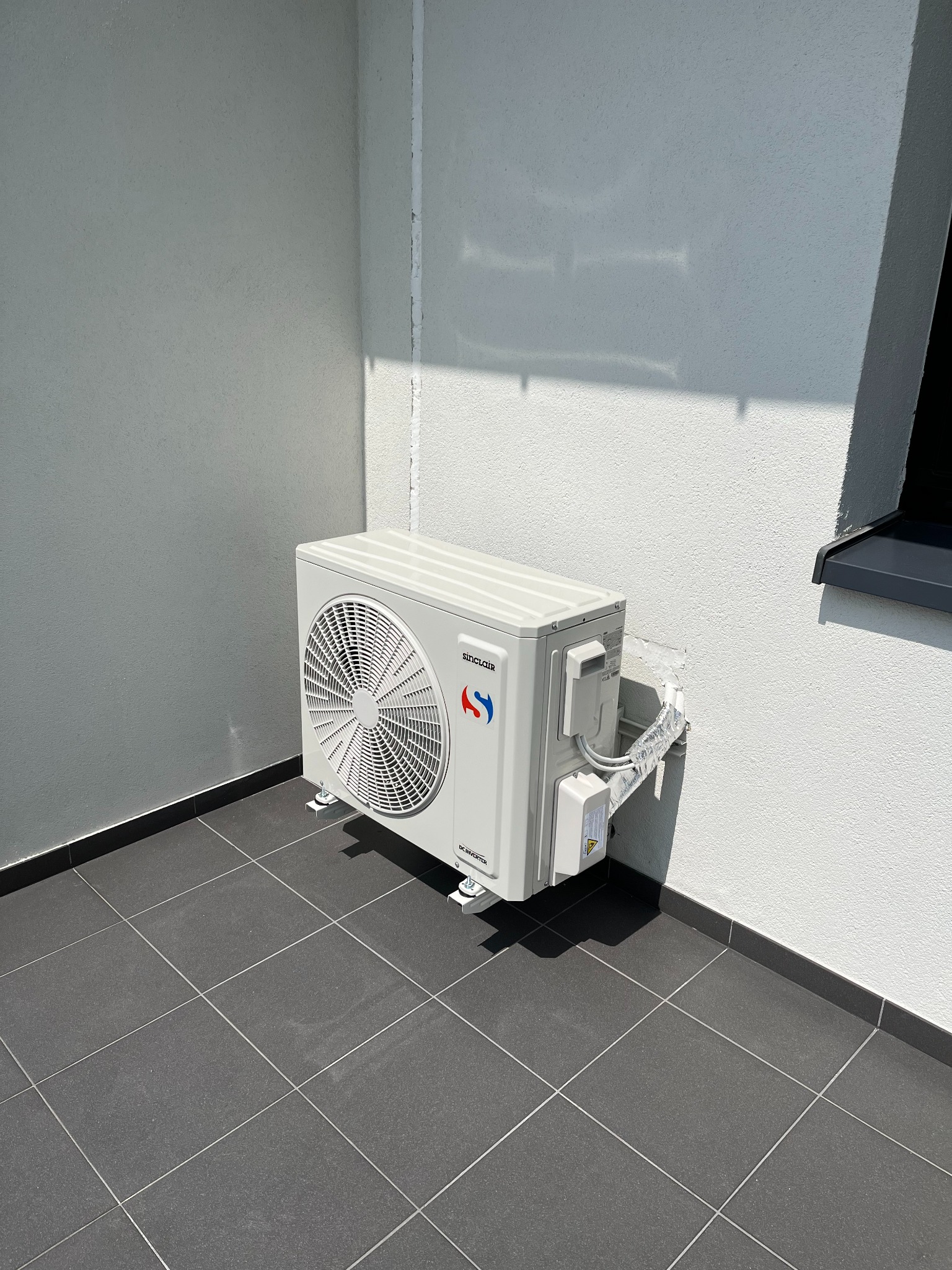 Biały klimatyzator zewnętrzny zamontowany na szarej, kafelkowanej podłodze przy białej ścianie budynku z widocznym oknem. Widoczne przewody w srebrnej osłonie.