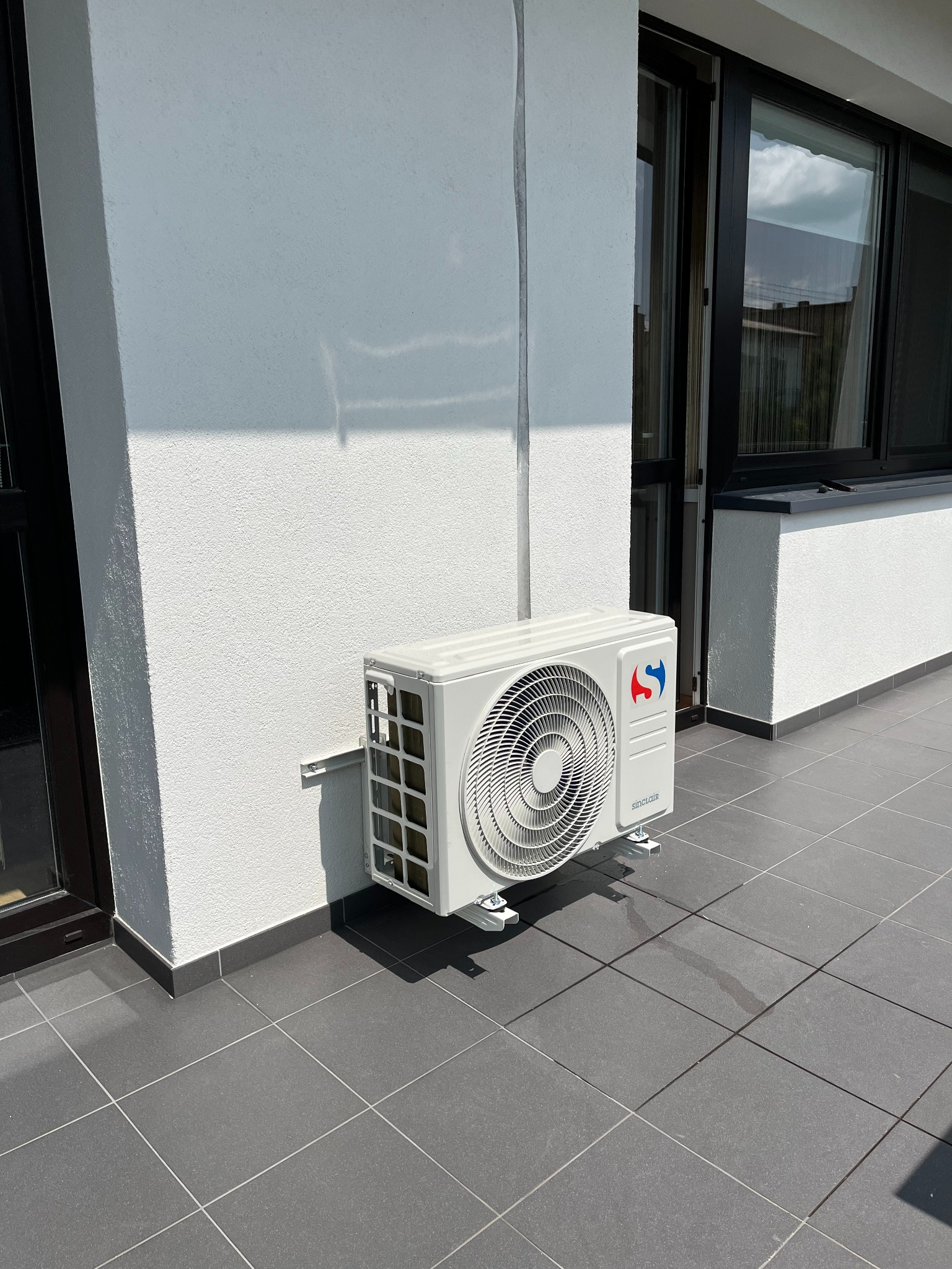 Biały klimatyzator Sinclair zamontowany na ścianie balkonu z szarymi płytkami, widoczne okna z czarnymi ramami i fragment elewacji budynku w słoneczny dzień.