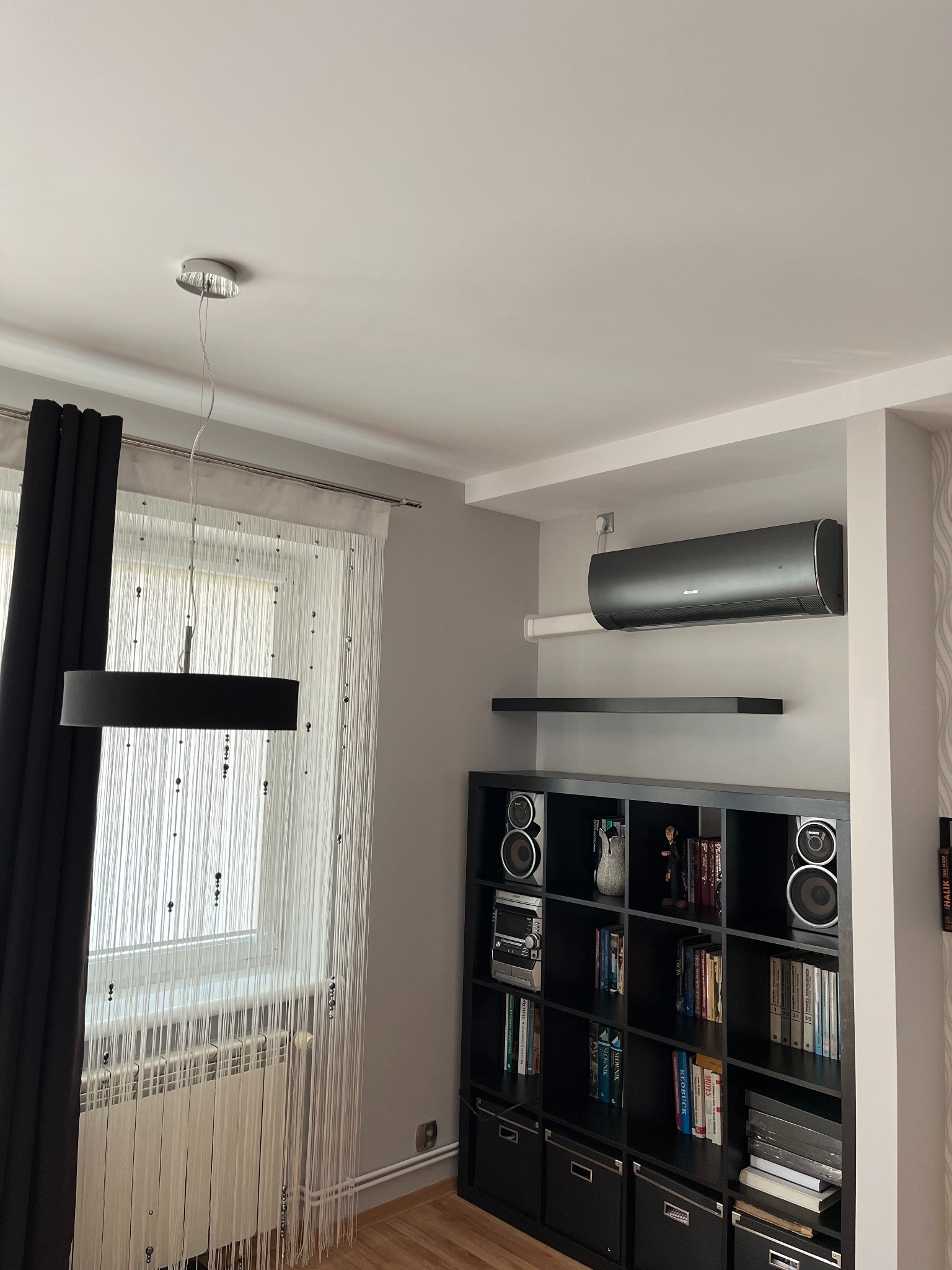 Czarny klimatyzator ścienny zamontowany nad czarnym regałem z książkami i sprzętem audio, oświetlony przez wiszącą lampę na tle okna z firankami.