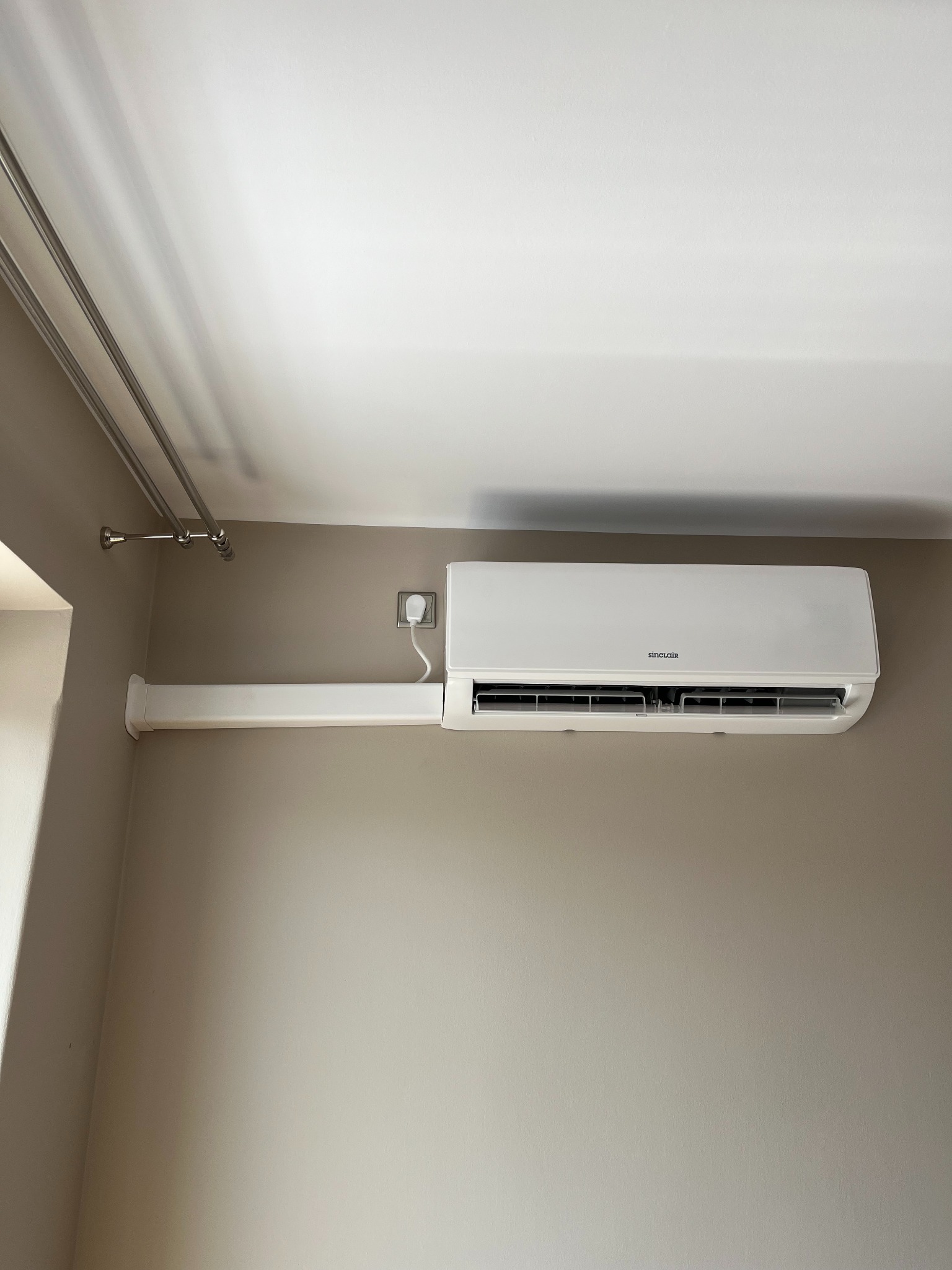 Biały klimatyzator Sinclair zamontowany na beżowej ścianie, z widoczną listwą maskującą przewody i gniazdkiem elektrycznym. W tle sufit i suszarka na pranie.