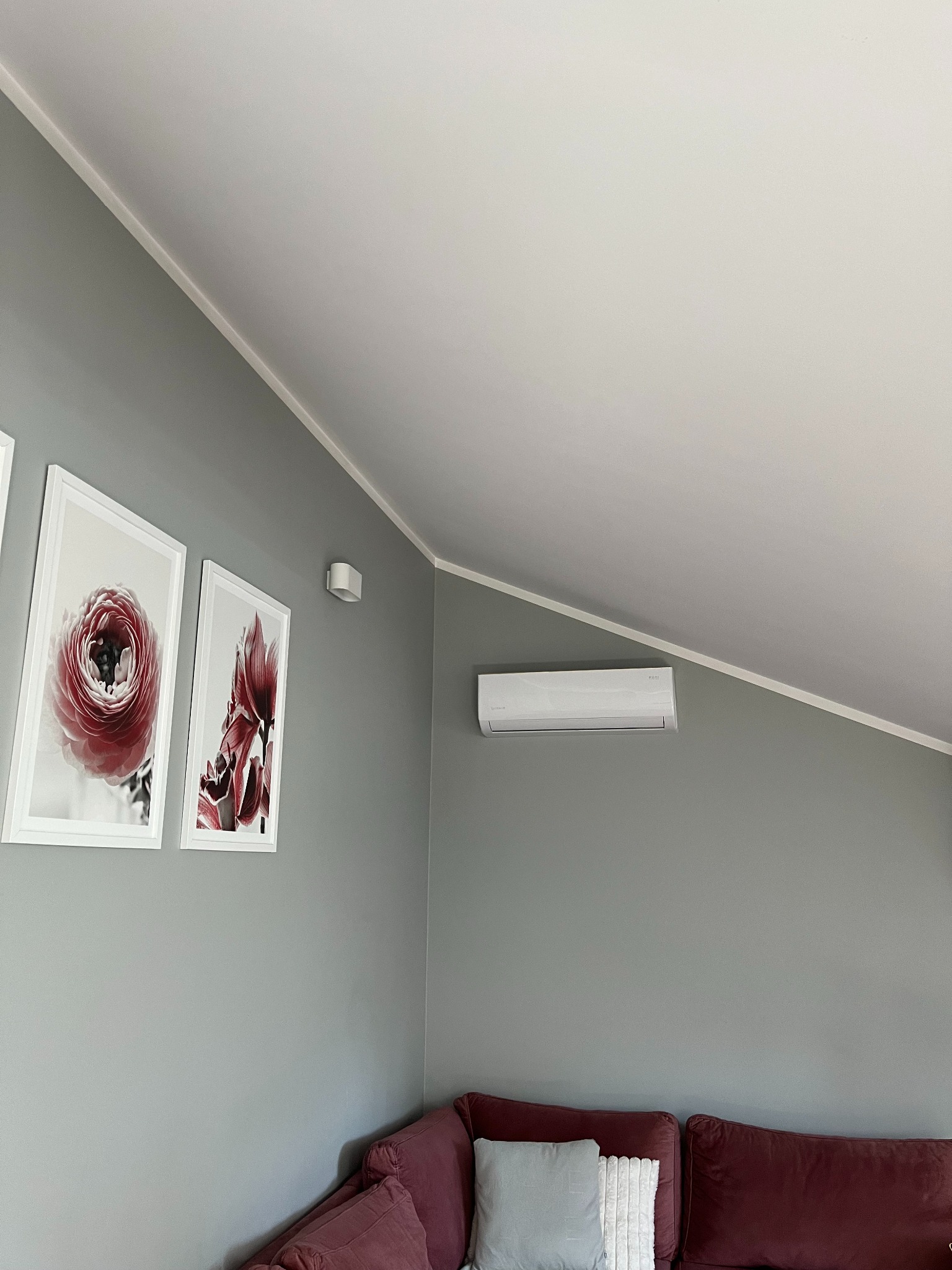 Biały klimatyzator ścienny w szarym pokoju z obrazami kwiatów i bordową sofą. Oświetlenie punktowe. Skośny sufit.