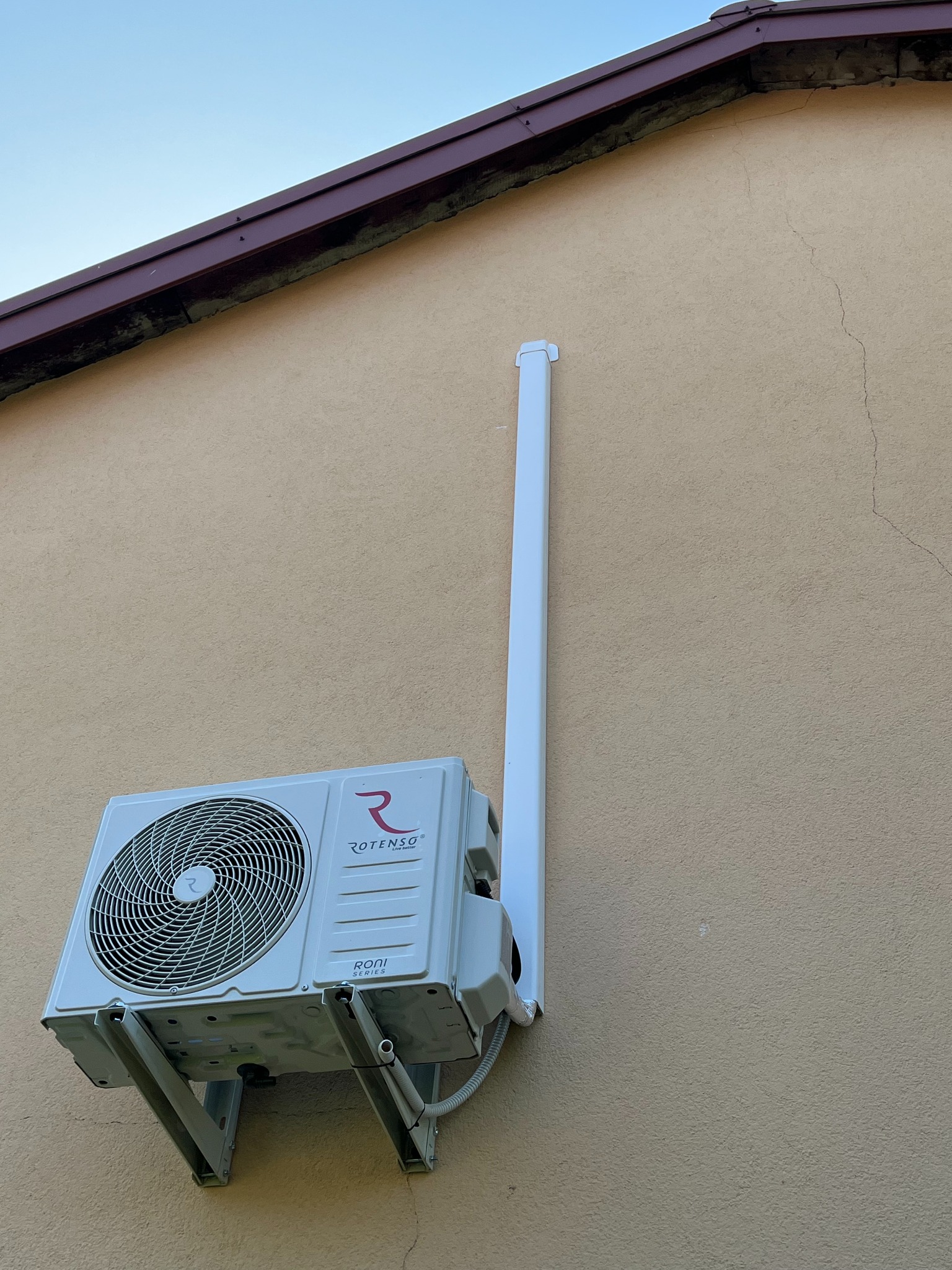 Zamontowana jednostka zewnętrzna klimatyzacji Rotenso Roni Series na elewacji budynku z widoczną rurą odprowadzającą skropliny. Ujęcie z dołu, jasne tło.