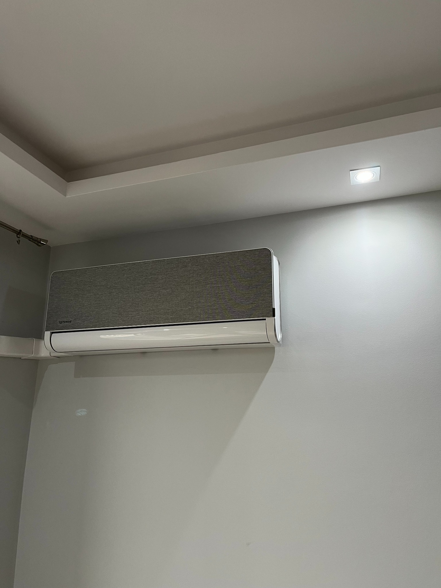Szary klimatyzator Rotenso zamontowany na ścianie w pomieszczeniu z oświetleniem punktowym. Widoczny fragment listwy przypodłogowej i karnisza.