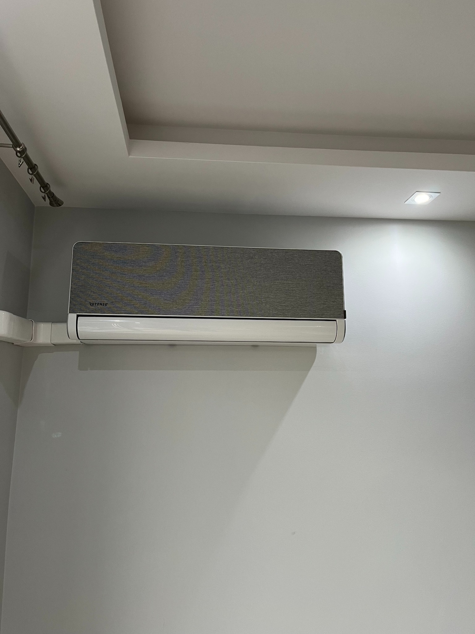 Szary klimatyzator Rotenso zamontowany na jasnej ścianie, widoczne białe rury instalacyjne i oświetlenie sufitowe. Minimalistyczny design, nowoczesne wnętrze.