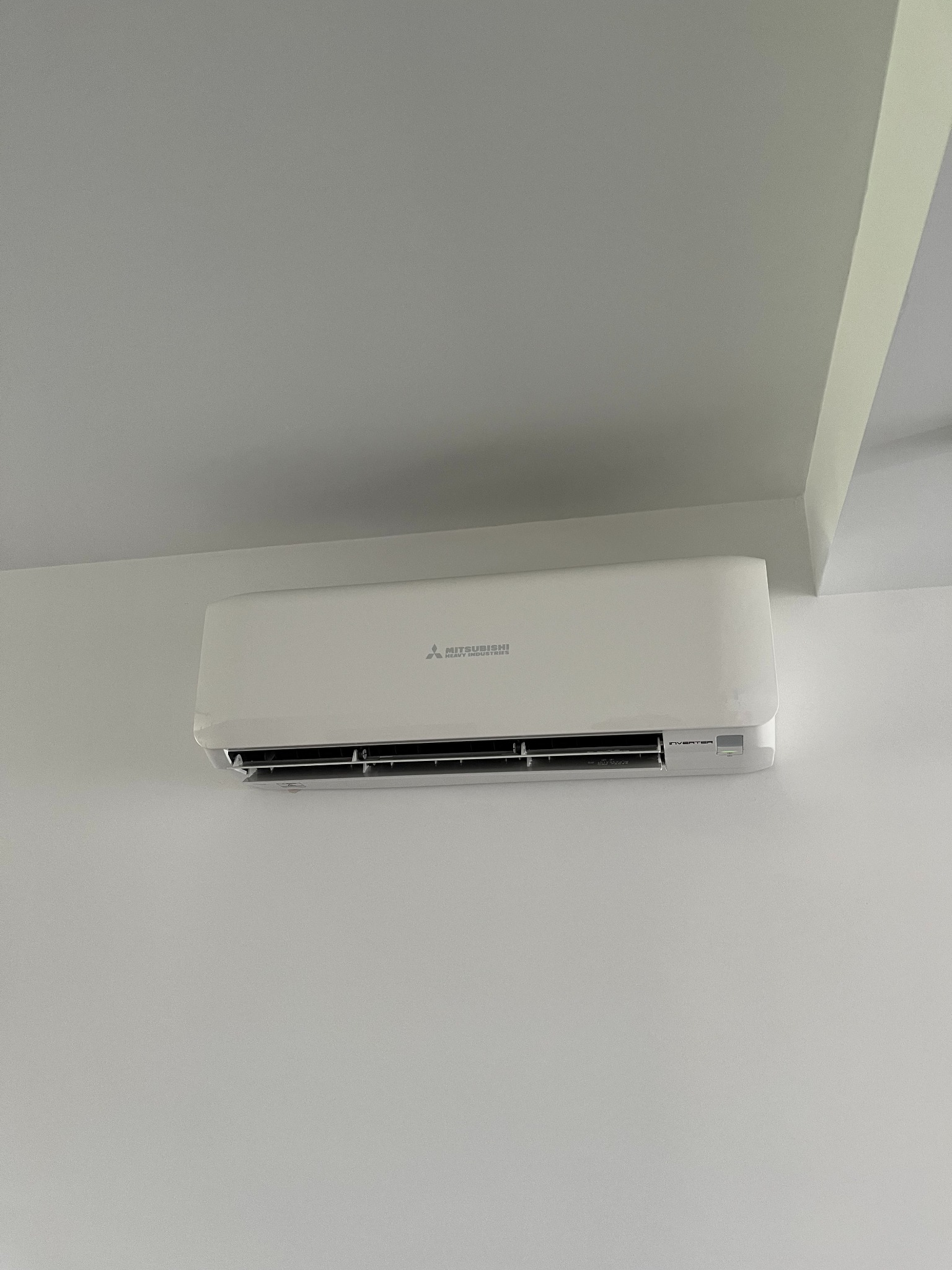 Biały klimatyzator Mitsubishi Electric zamontowany na ścianie pod sufitem w jasnym pomieszczeniu. Widoczny z bliska, z delikatnym oświetleniem.