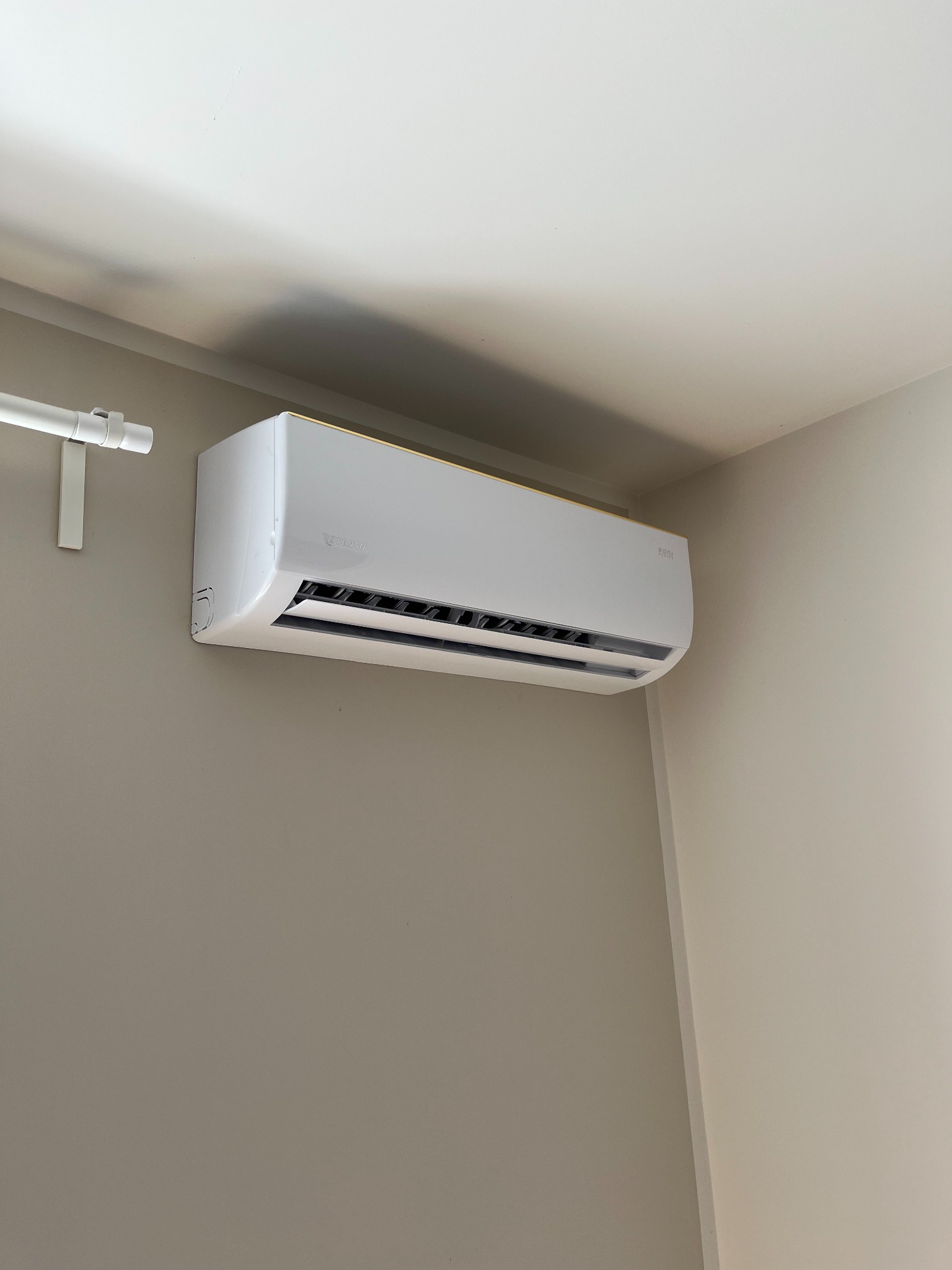 Biały klimatyzator Rotenso zamontowany na beżowej ścianie w rogu pomieszczenia, widoczny fragment karnisza. Minimalistyczny design, nowoczesne rozwiązanie chłodzące.