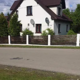 Nowoczesne, poziome ogrodzenie z ciemnego drewna z białymi słupkami na tle białego domu z brązowym dachem i dwoma antenami satelitarnymi.