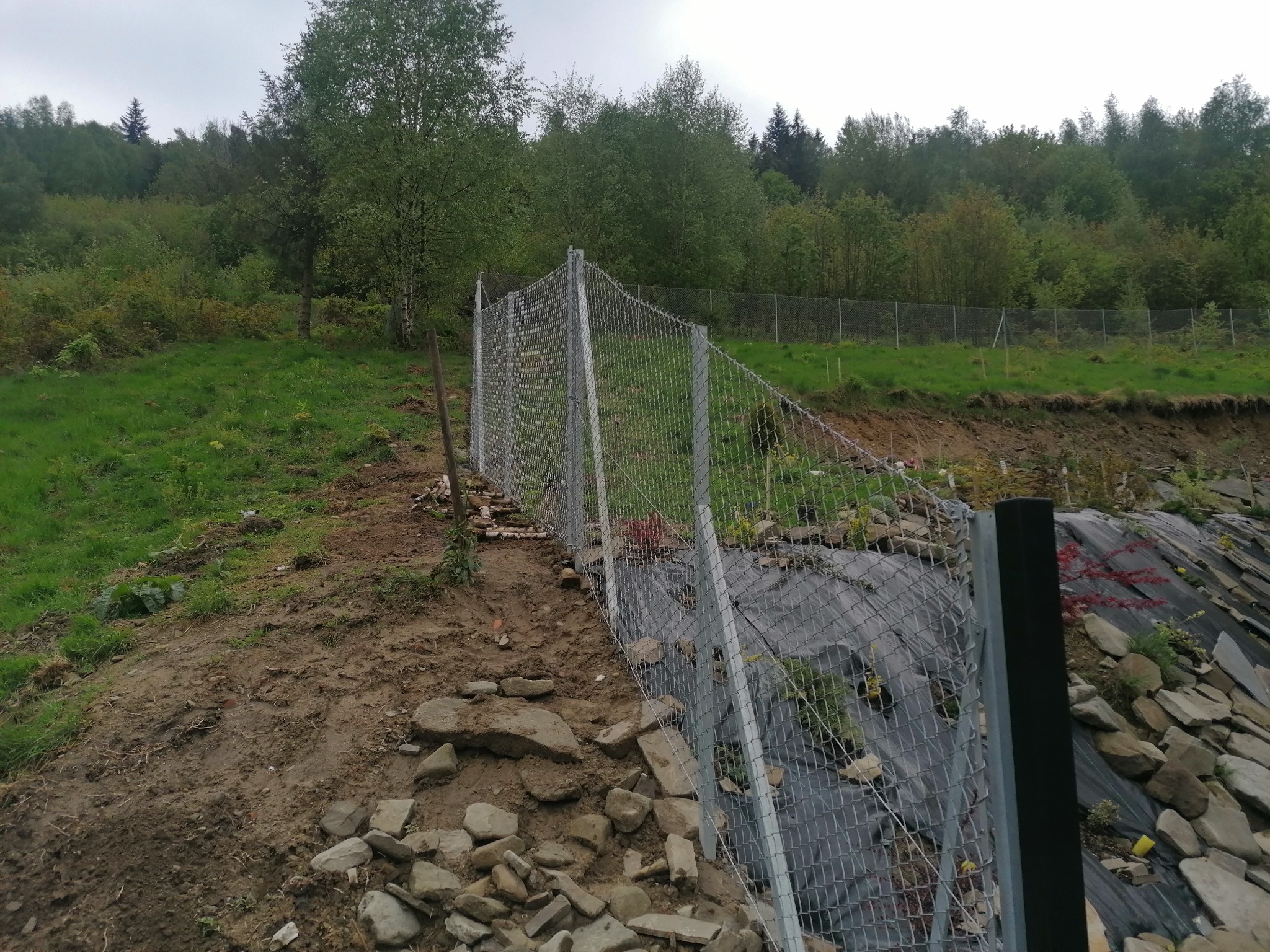 Metalowa siatka ogrodzeniowa na zboczu wzgórza, zabezpieczająca teren z kamieniami i czarną folią, w tle drzewa.