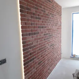 Ściana wykończona czerwoną cegłą klinkierową z podświetleniem LED wzdłuż krawędzi, widoczne gniazdka elektryczne na sąsiedniej ścianie, surowe wykończenie podłogi.